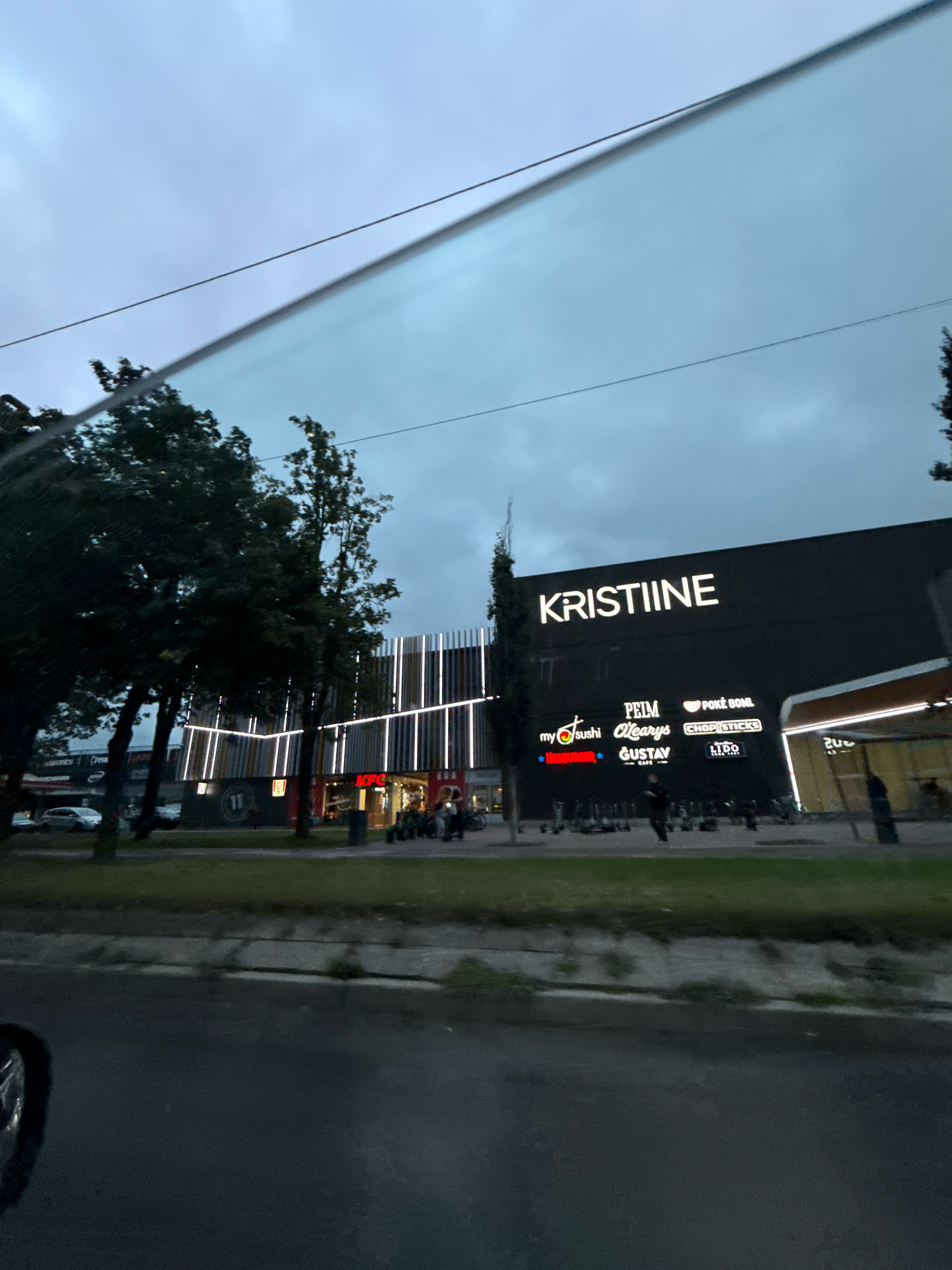 The image shows a modern shopping center exterior with the prominent "KRISTIINE" sign illuminated in white. Below the main sign, several smaller illuminated signs advertise businesses such as "my sushi," "PEIM," "O'Learys," "POKE BOWL," "CHOPSTICKS," and "GUSTAV CAFE." The shopping center features vertical white lighting accents. In front of the building, a line of electric scooters is parked. To the left of the shopping center, trees are visible, and a street with cars is present. The sky is overcast. The scene depicts an urban commercial area, specifically the Kristiine shopping center in Tallinn, Estonia.