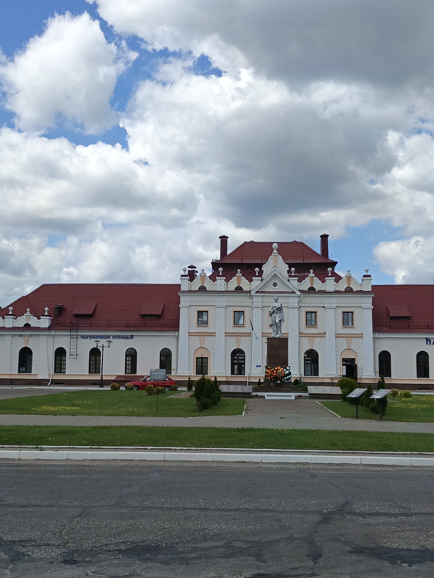The image captures a frontal view of a two-story train station building with a distinctive red tiled roof, located in the city of Парэчча, Belarus. The building's facade is cream-colored with beige decorative elements around the windows and cornices. A statue of a soldier stands prominently in front of the central section of the building, positioned on a stone pedestal adorned with a wreath of flowers. The area in front of the station is paved with gray bricks and surrounded by green grass. A red car is parked on the left side of the building, and a street lamp is visible between the car and the entrance. The sky above is filled with a mix of white and gray clouds. The scene appears to be an outdoor, public space, likely a train station plaza.