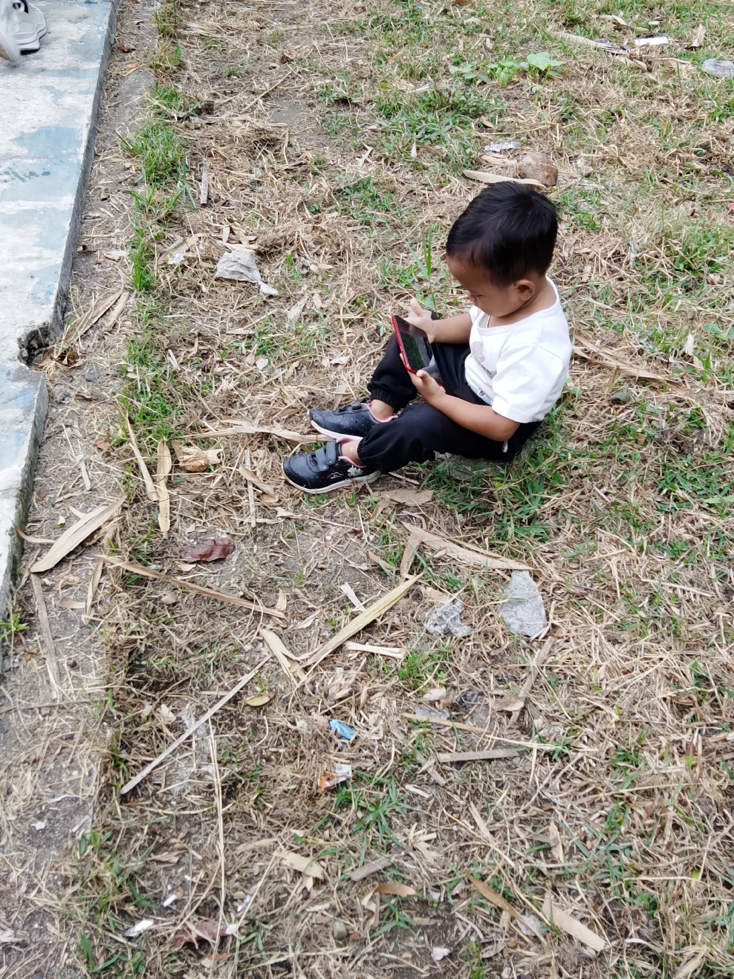A young child sits on the ground in Jumo, Indonesia, engrossed in a red smartphone. The child is wearing a white t-shirt and black pants with black sneakers. The ground is covered in dry grass, fallen leaves, and some scattered debris. To the left of the child is a concrete structure with a blue painted edge. The scene appears to be outdoors, possibly in a park or a less maintained area.