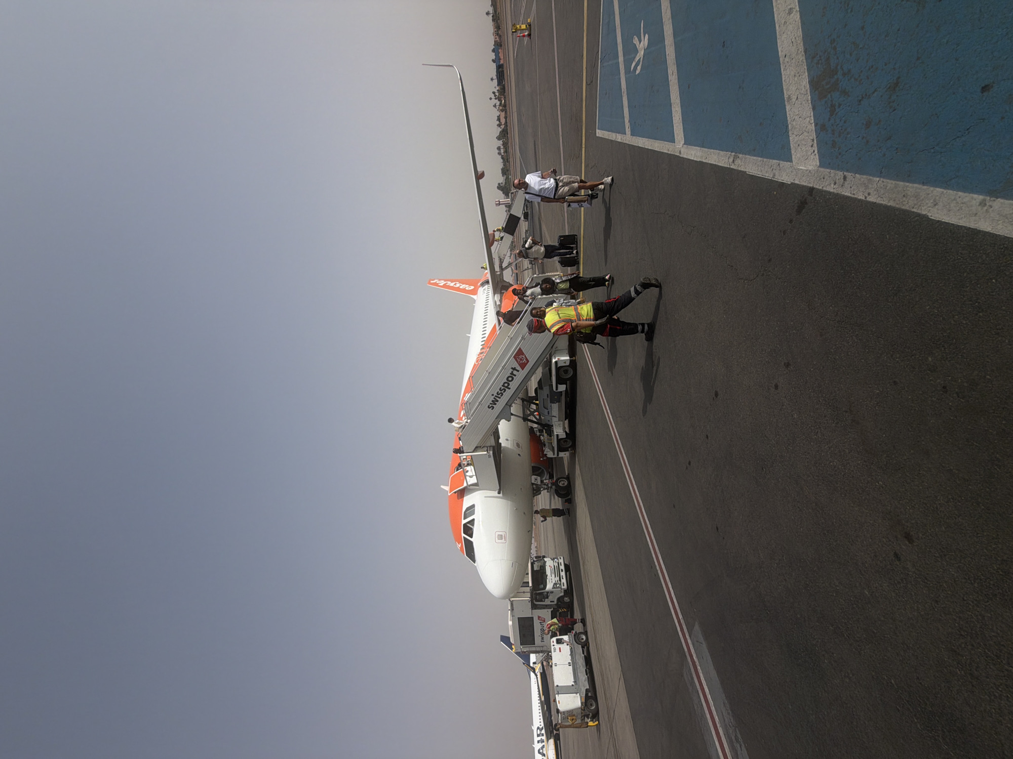 An EasyJet airplane is parked at an airport gate. A mobile stairway is attached to the front left passenger door, and several people, including passengers and ground crew, are on the stairs and near the aircraft. A man in a high-visibility vest is walking away from the plane. To the left of the EasyJet plane is a Ryanair airplane. Ground service vehicles are positioned around the EasyJet aircraft. The scene is on an asphalt tarmac under a clear sky. The image appears to be taken at Marrakesh Menara Airport in Marrakesh, Morocco, based on the EasyJet branding and the presence of palm trees in the background, characteristic of the Moroccan landscape.