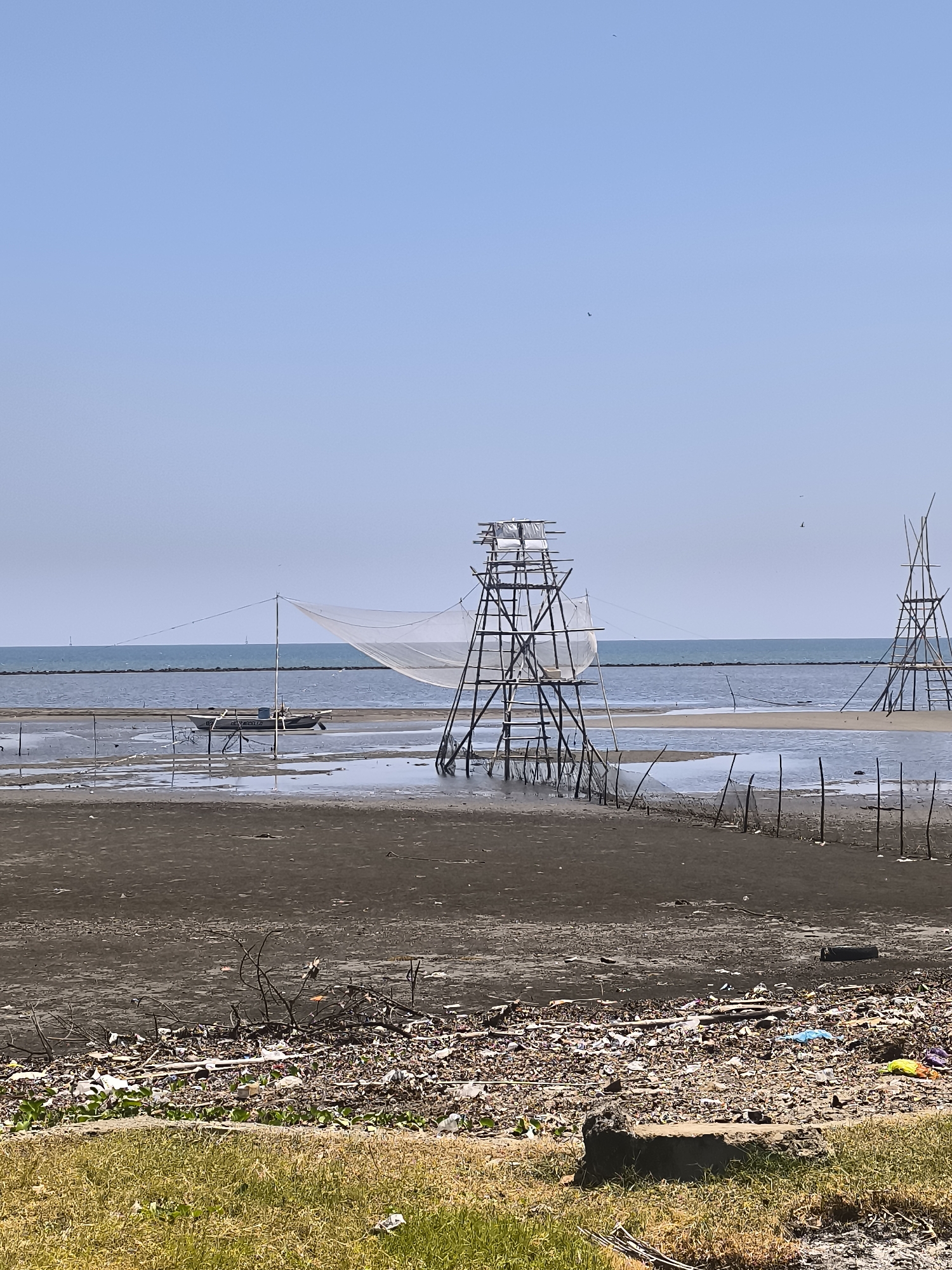 This image depicts a coastal scene in Barru, Indonesia, characterized by a tidal flat exposed under a clear blue sky. In the foreground, a strip of dry, green grass is visible, bordered by a large accumulation of debris, including plastic waste, branches, and organic material. The tidal flat itself is dark and muddy, with shallow pools of water reflecting the sky.
Dominating the mid-ground are two wooden fishing structures. The one on the left is a tall, multi-tiered platform made of bamboo or similar wood, supporting a large white fishing net stretched between poles. A small boat, appearing to be a traditional fishing vessel, is moored in the shallow water near this structure. The second fishing structure, on the right, is also a tall wooden frame, though less elaborate and without a visible net. A line of wooden stakes stretches across the tidal flat, possibly marking boundaries or part of a fishing apparatus.
The background shows the calm, blue sea meeting the horizon, with a faint line of what appears to be a distant breakwater or a line of land. The sky is clear, with only a few very small, dark specks that could be birds. The overall impression is of a functional, albeit somewhat desolate, fishing area with evidence of significant litter accumulation along the shoreline.