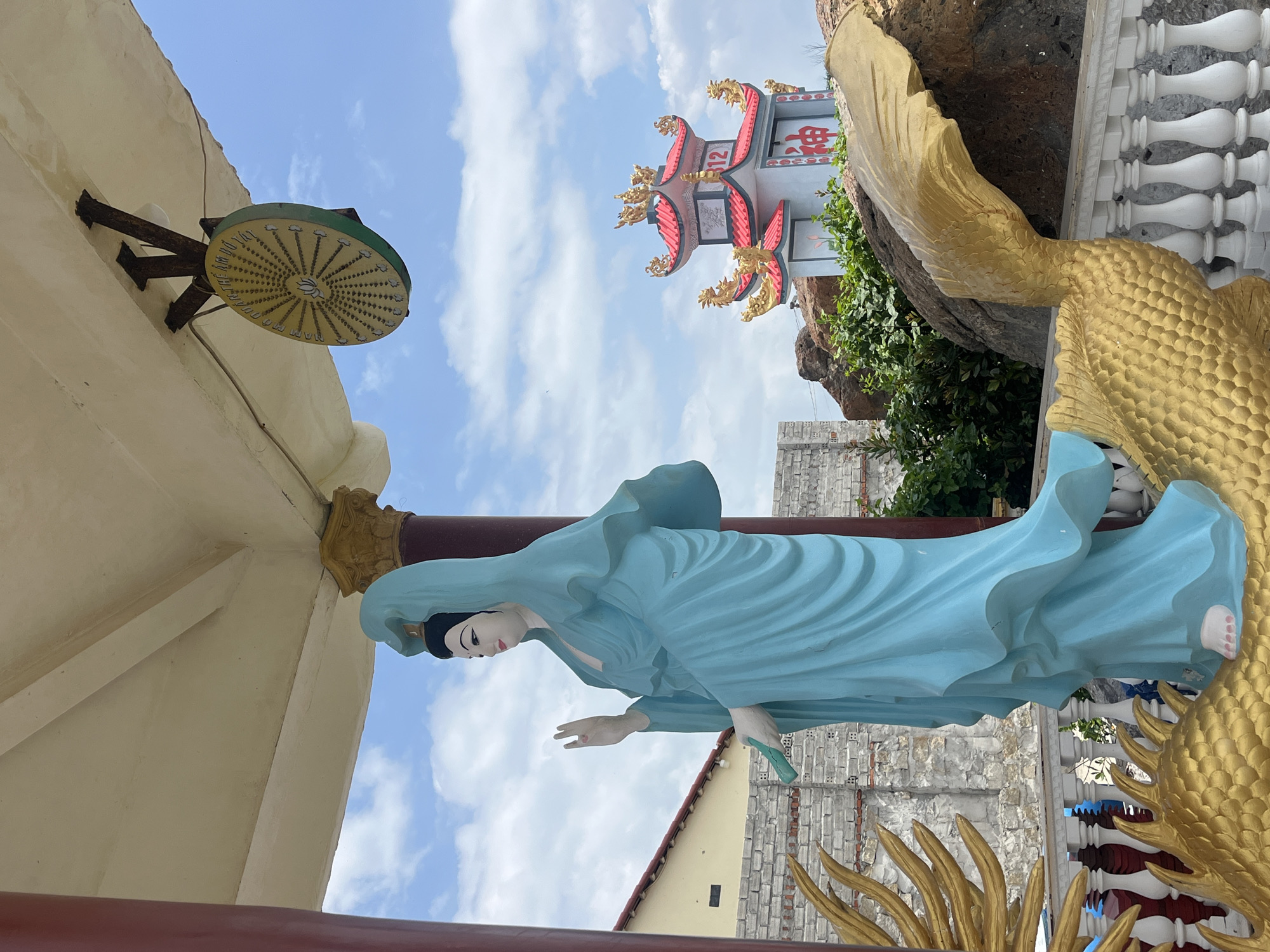 A serene statue of Guanyin, the Bodhisattva of Compassion, stands prominently outdoors in Mui Ne, Vietnam. The statue, rendered in a tranquil blue, is depicted in a flowing robe, with one hand raised in a gesture of blessing and the other holding a green object. It rests upon a large, golden, scaly mythical creature, possibly a dragon or phoenix, which curves around the base.
Behind Guanyin, a traditional Vietnamese pagoda-style building rises against a bright blue sky dotted with white clouds. This structure features ornate red and gold roofing, elaborate carvings, and the number "312" displayed prominently. The surrounding environment includes textured brick walls, rocky outcrops, and lush green foliage, suggesting a temple or spiritual site within the coastal city of Mui Ne. A circular yellow emblem with intricate patterns and a lotus flower at its center hangs from the overhang of a covered structure to the left of the pagoda. The overall scene conveys a sense of peace and cultural richness.