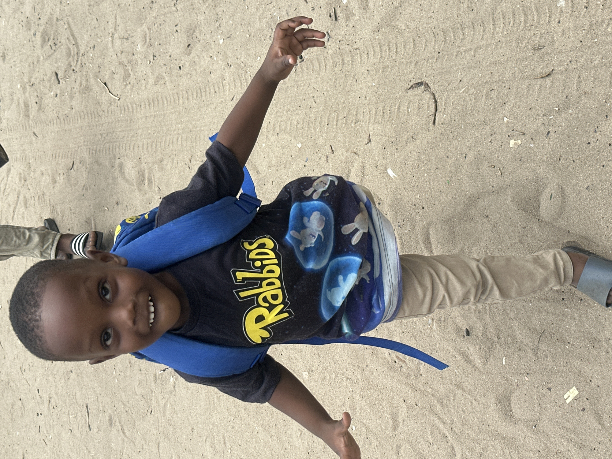 A young boy, likely of African descent, is pictured standing on a sandy surface, possibly a beach or a dirt road, in Badagry, Nigeria. He is wearing a dark t-shirt with a "Rabbids" logo and cartoon images, tan pants, and grey sandals. A blue backpack is slung over his shoulders. His left leg is slightly raised, suggesting he is in motion. The background consists of sand with tire tracks and scattered debris. The lighting suggests it is daytime.