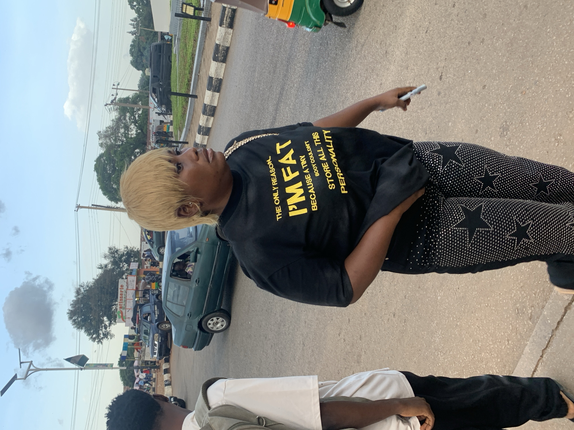 The image is taken at a street intersection in Jos, Nigeria. In the foreground, a woman with blonde hair is walking and looking to her right. She is wearing a black t-shirt with yellow text that reads "THE ONLY REASON I'M FAT BECAUSE A TINY BOOY COULDN'T STORE ALL THIS PERSONALITY". She is also wearing black pants adorned with numerous rhinestones and star shapes. To her left, another person is partially visible, wearing a white t-shirt and a backpack.
In the background, there is a street with several vehicles, including a green sedan, a blue police pickup truck, and a black van. There are also buildings, traffic lights, and overhead power lines. A small, colorful auto-rickshaw is visible on the right side of the image, partially obscuring the woman. The sky is partly cloudy. The overall scene depicts a busy street corner in an urban environment.