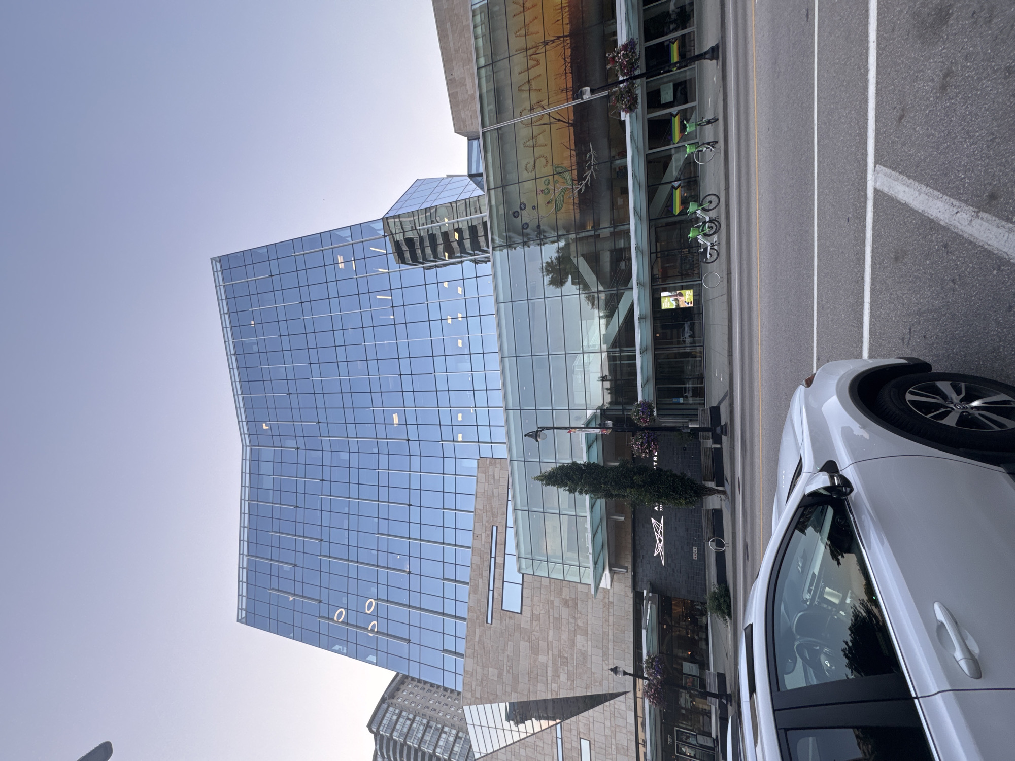 The image shows the exterior of the Anvil Centre in New Westminster, Canada, on a clear day. A white SUV is parked in the foreground, facing away from the camera. Beyond the car is a street with a sidewalk and several electric bikes parked in a rack. The Anvil Centre is a modern building with a glass facade that reflects the sky and surrounding buildings. The building has a distinctive curved roofline. The street level of the building features a row of businesses. The sky is a pale blue, and the overall lighting suggests it is either early morning or late afternoon.