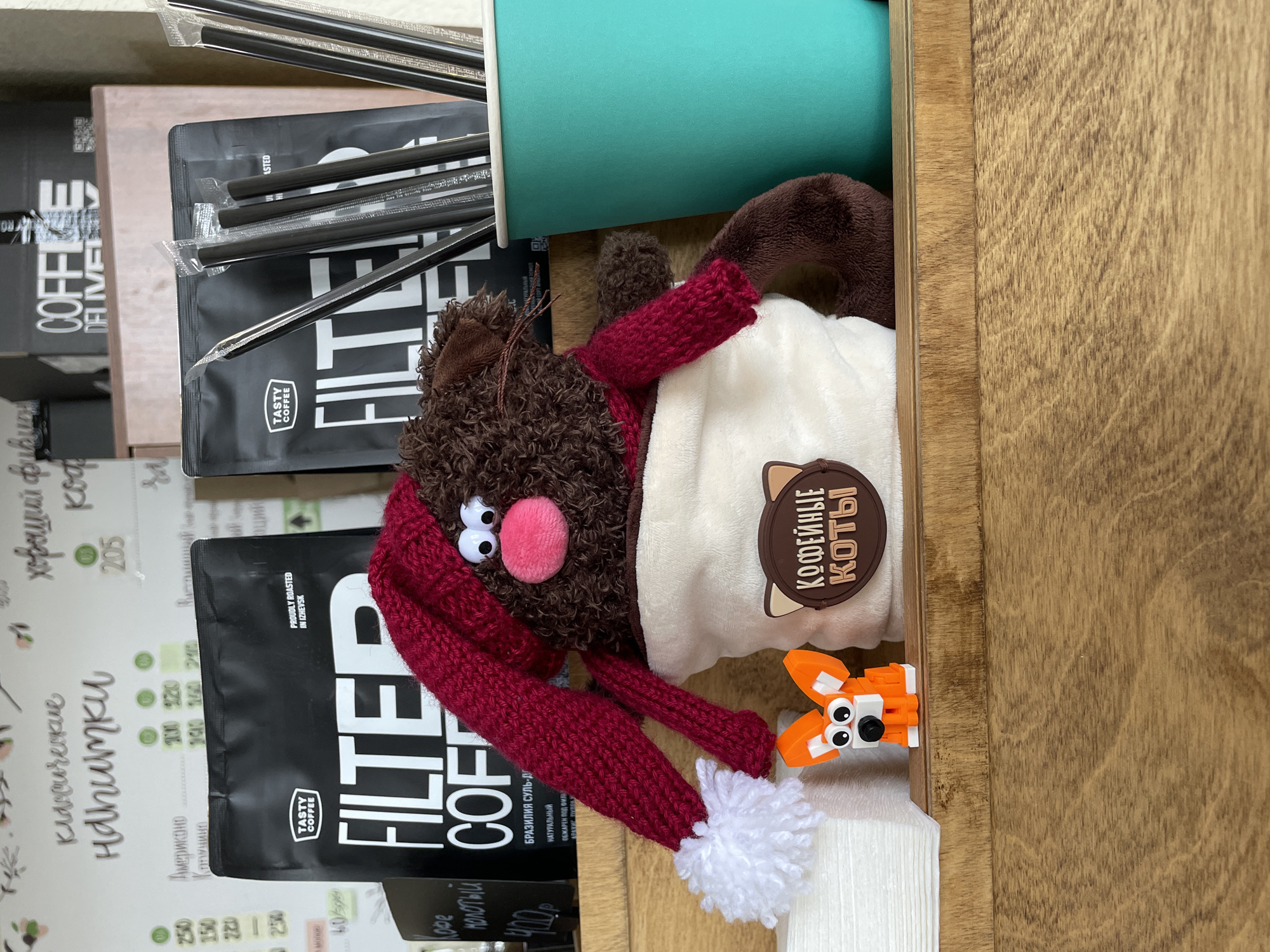 A vertically oriented close-up shot shows a section of a shelf or display. On the shelf are packets of coffee beans, individually wrapped straws, a blue cup, and stuffed animals.
Specifically, there are at least two packets of “Tasty Coffee Filter Coffee” from Izhevsk, Russia on the left, with nutritional information partially visible in Cyrillic script. The packets are black and white. There is a bundle of straws wrapped in clear plastic packaging behind the top packet of coffee. A light blue cup sits behind the bundle of straws and to the right.
A brown stuffed animal with a pink nose and a red knitted hat is nestled among the items, along with a white stuffed pillow bearing a logo in the shape of a cat head that says "Кофейные Коты" in Cyrillic script. An orange brick-built animal figurine (possibly a fox) is positioned near the edge of the shelf.