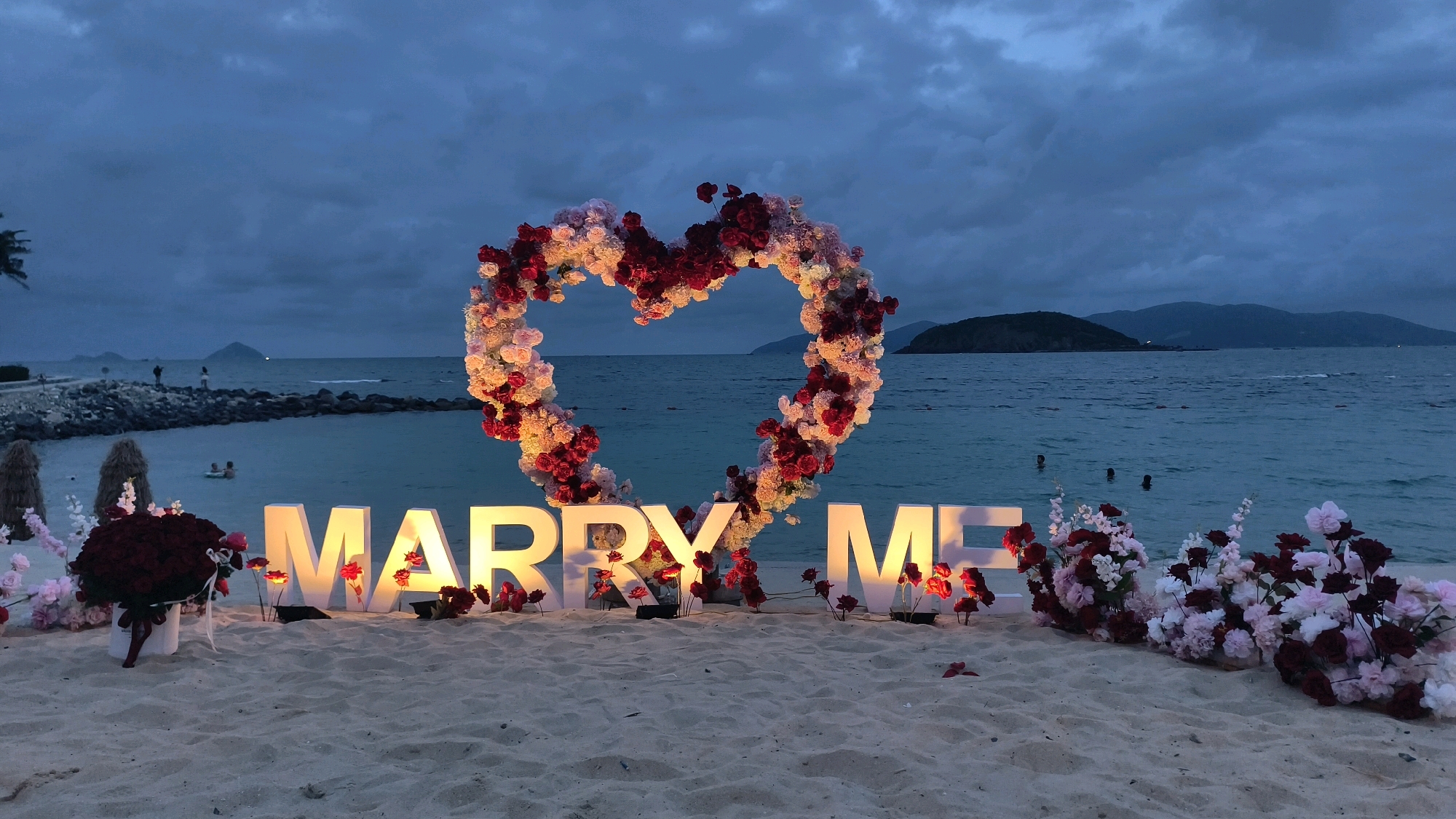 The image depicts a romantic proposal setup on a sandy beach at dusk. In the foreground, illuminated white block letters spell out "MARRY ME". These letters are adorned with flowers, and a large heart-shaped floral arrangement, predominantly in shades of pink and deep red, is positioned behind them, framing the "ME" of the message. To the left and right of the letters are bouquets of flowers and additional floral decorations scattered on the sand. The beach stretches towards the ocean, which is calm and blue, with a distant coastline and hills visible under a cloudy, twilight sky. Several people are in the water, appearing to swim or wade. The overall ambiance is serene and suggestive of a special occasion.