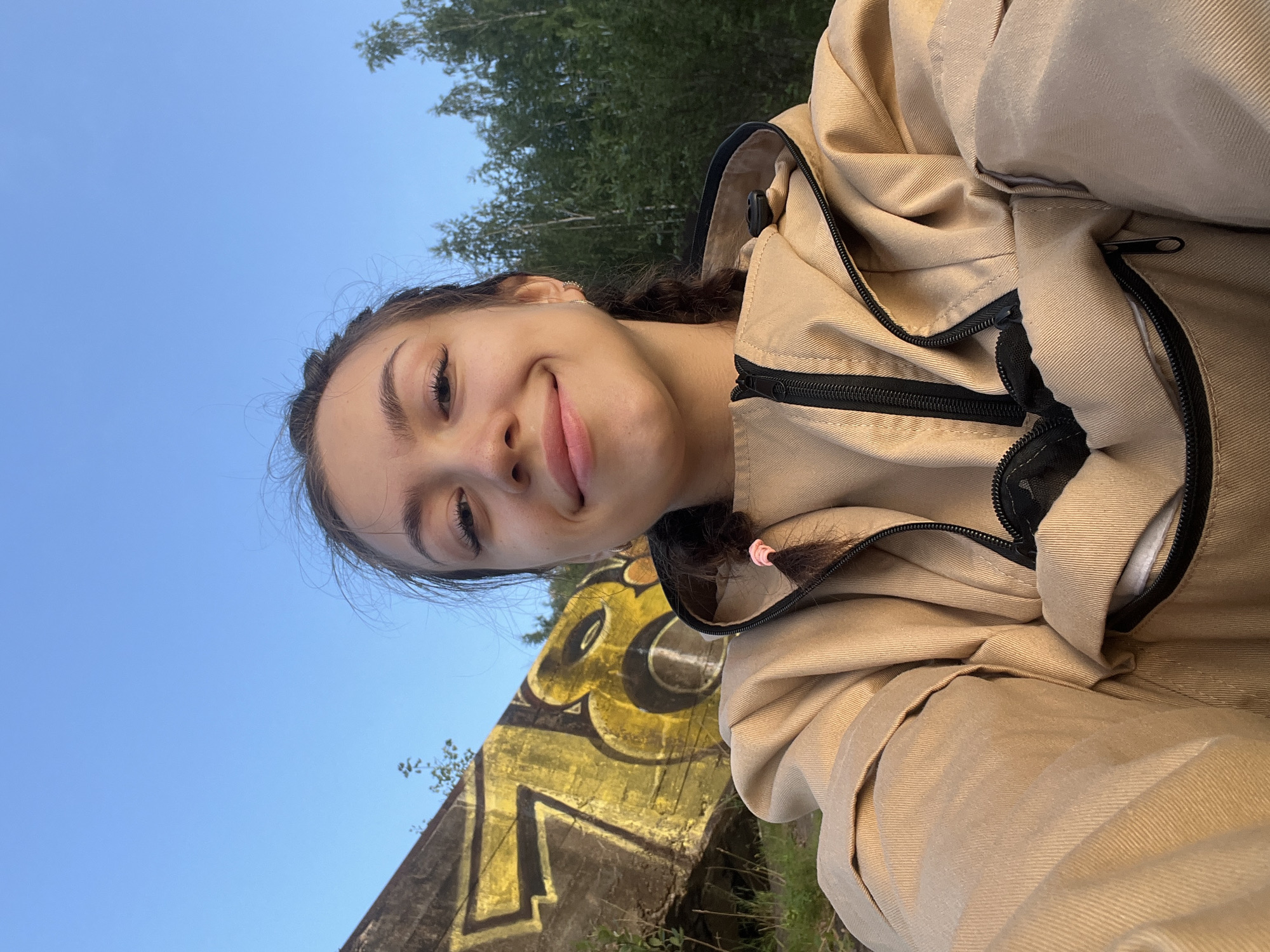 A young woman with dark hair in braids, wearing a tan jacket, smiles at the camera. She is outdoors, with a blue sky and trees in the background. To her left, a concrete structure with yellow graffiti is visible. The scene appears to be in Chernushka, Russia.