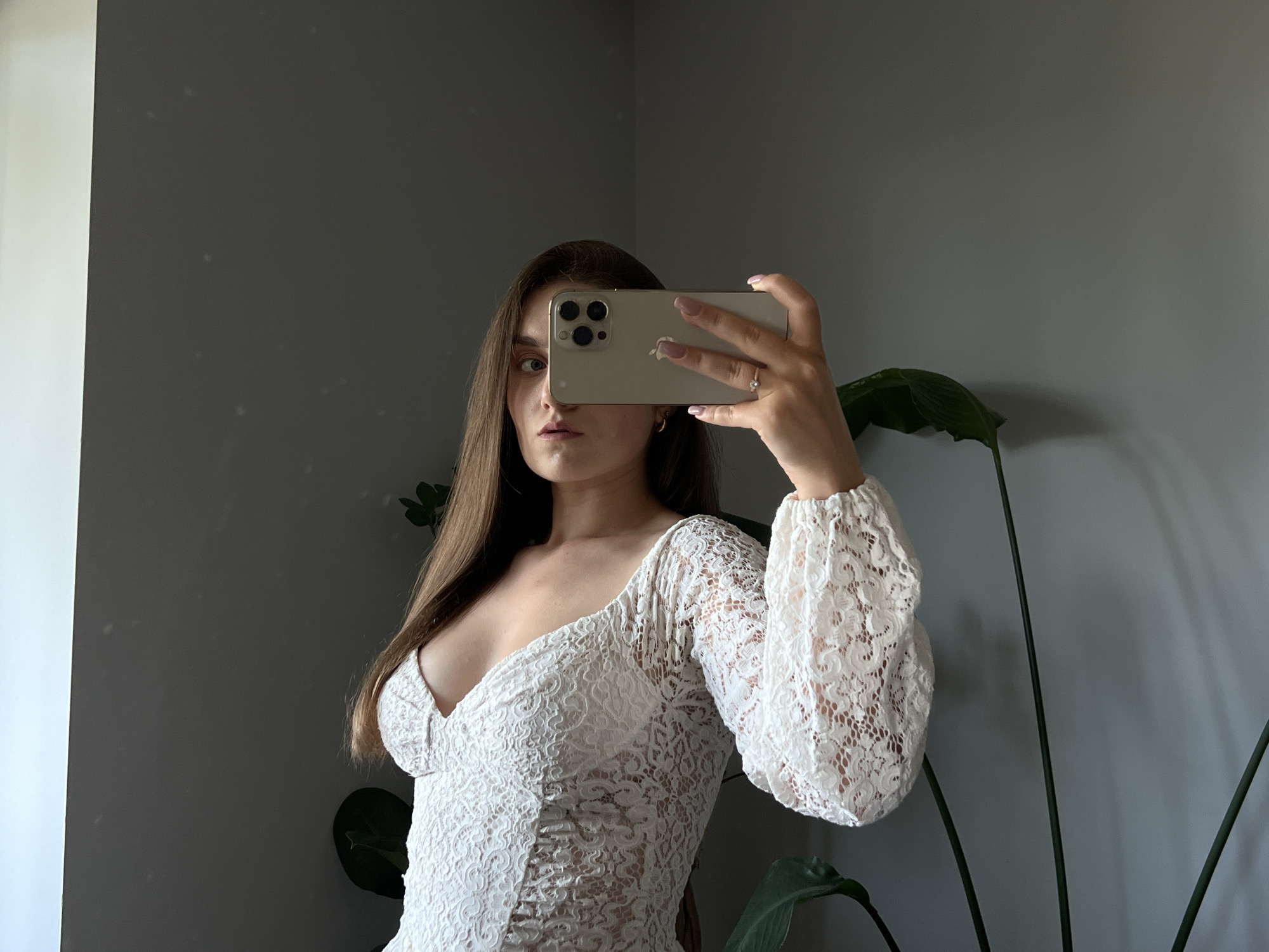 The media file is a medium shot, taken indoors, of a young woman with long, dark blonde hair. She is holding a smartphone in her right hand, covering the top half of her face, and taking a selfie in a mirror. She is wearing a white, lace dress with a deep V-neck and long, puffed sleeves. The dress has ruching on the front bodice.
The background is a plain, gray wall. To the left, a sliver of a white wall or object is visible. To the right, a large, green plant with broad leaves is partially in frame. The lighting suggests it is daytime, possibly late afternoon due to the soft shadows. The mood appears neutral to slightly inquisitive, with the woman's visible eye looking directly at the camera. There is no visible text in the image. The location context is Ternopil, Ukraine.