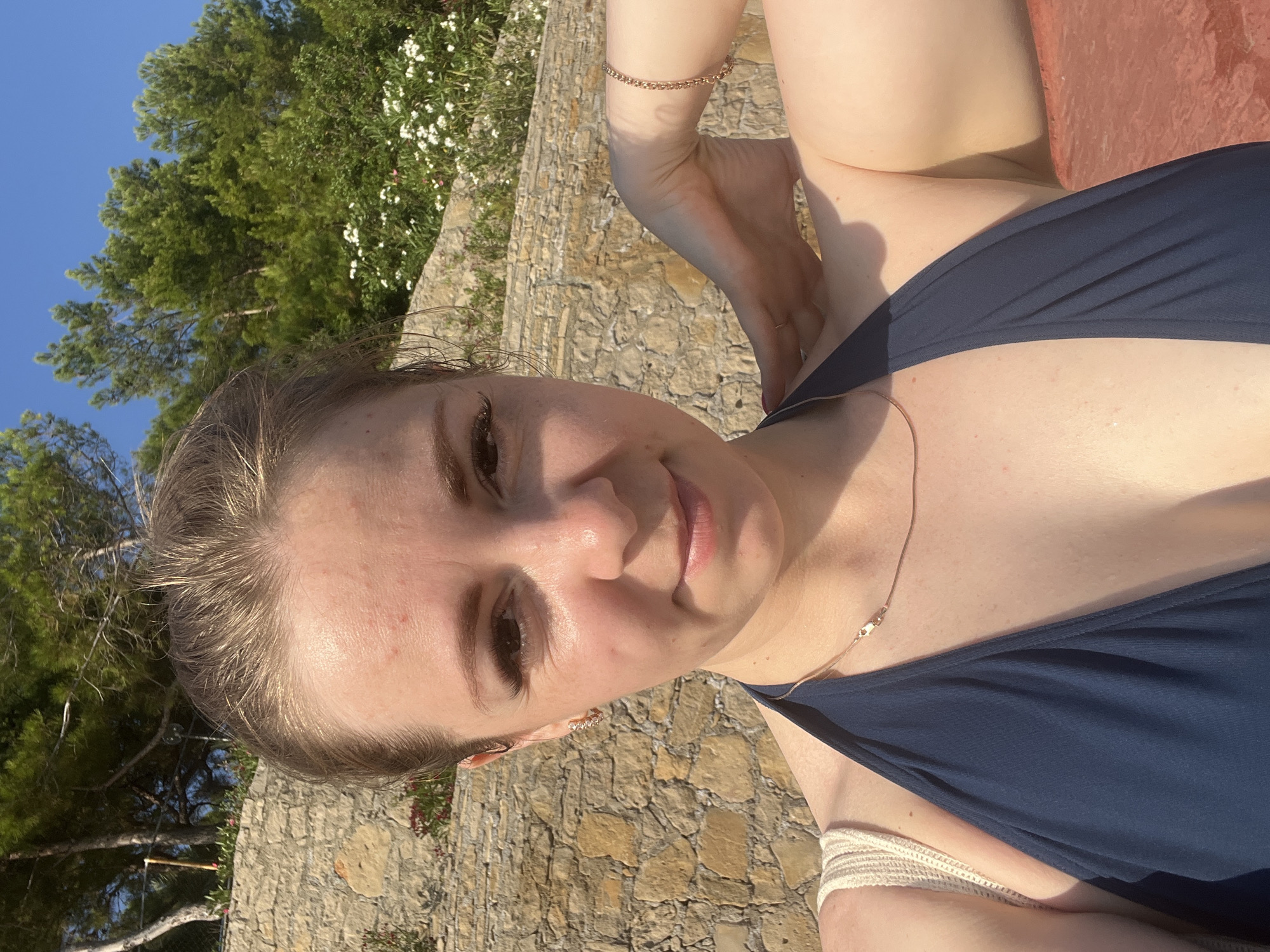A close-up, head-and-shoulders selfie taken outdoors shows a young woman with blonde hair pulled back, smiling slightly at the camera. She is wearing a dark blue halterneck swimsuit and a thin gold necklace. A gold bracelet is visible on her right arm, which is bent and resting on her neck. Her left shoulder is also visible, with a portion of a straw bag strap.
The background features a rustic stone wall, abundant green foliage, and white flowers, suggesting a natural or garden setting. The bright sunlight and blue sky indicate it is daytime. The presence of pine trees and Mediterranean flora suggests the location could be the Alanya peninsula in Turkey, possibly on a terrace or hillside overlooking the sea.