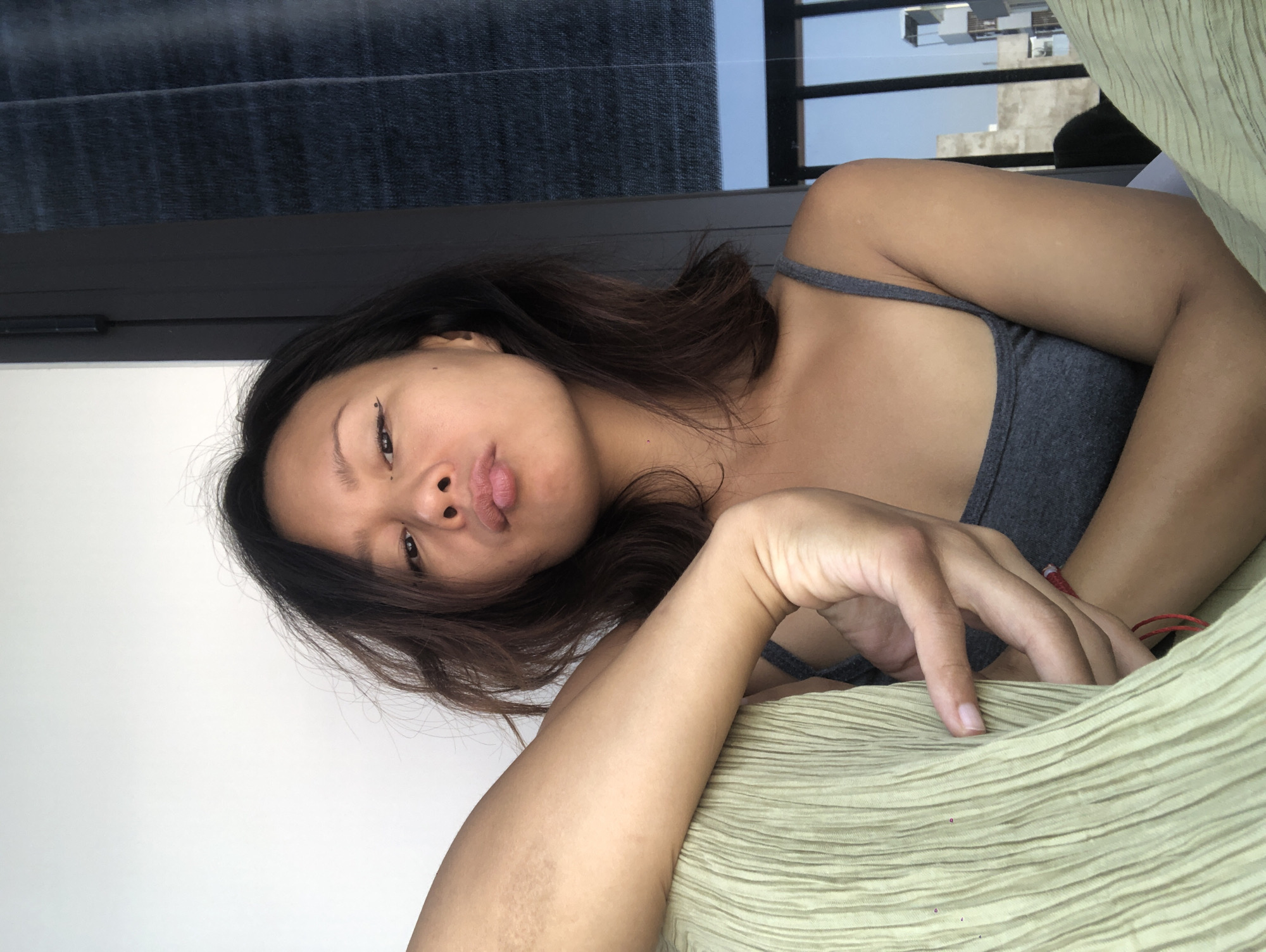 The image captures a young Asian woman lying down indoors, likely in Đà Nẵng, Vietnam, during the daytime. She is positioned with her head slightly tilted to her left, looking directly at the camera with a relaxed and slightly pouting expression. Her dark, medium-length hair cascades around her face and shoulders. She is wearing a grey tank top or camisole.
Notable details on her face include expressive eyes and full lips. Small black dots, possibly decorative eyeliner, are visible near the outer corner of her left eye and below her right eye. Her left arm is bent, with her hand and forearm resting on a light green, horizontally textured fabric, which appears to be a pillow or blanket. Her right hand is also visible, with fingers resting on the same fabric, and a thin red string (possibly a bracelet) is tied around her right wrist. Some skin discoloration or texture is visible on her left forearm.
The background behind her head is a plain white wall. To her right, a dark horizontal window frame is visible, with dark blinds or curtains above it. Through the window, a bright blue sky and distant urban buildings or structures can be seen, indicating an outdoor city view. The overall mood is casual and intimate. There is no visible text in the image.