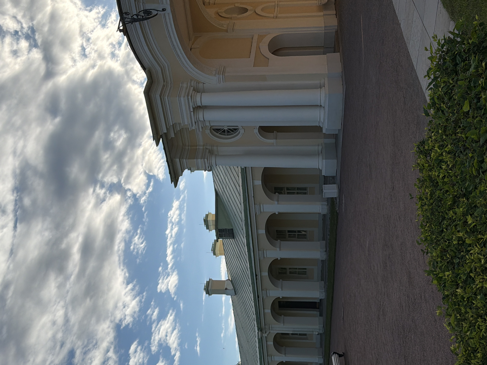 An exterior, low-angle, full shot features a long, yellow, classical-style building with a green metal roof under a partly cloudy blue sky. The building has a series of arches supported by white columns that lead to entrances and windows. The central part of the building features a grand arched entrance with decorative molding and additional white columns. The ground in front of the building is covered in dark gravel. In the foreground, there is a well-maintained green hedge. The scene appears to be a historical landmark or park.