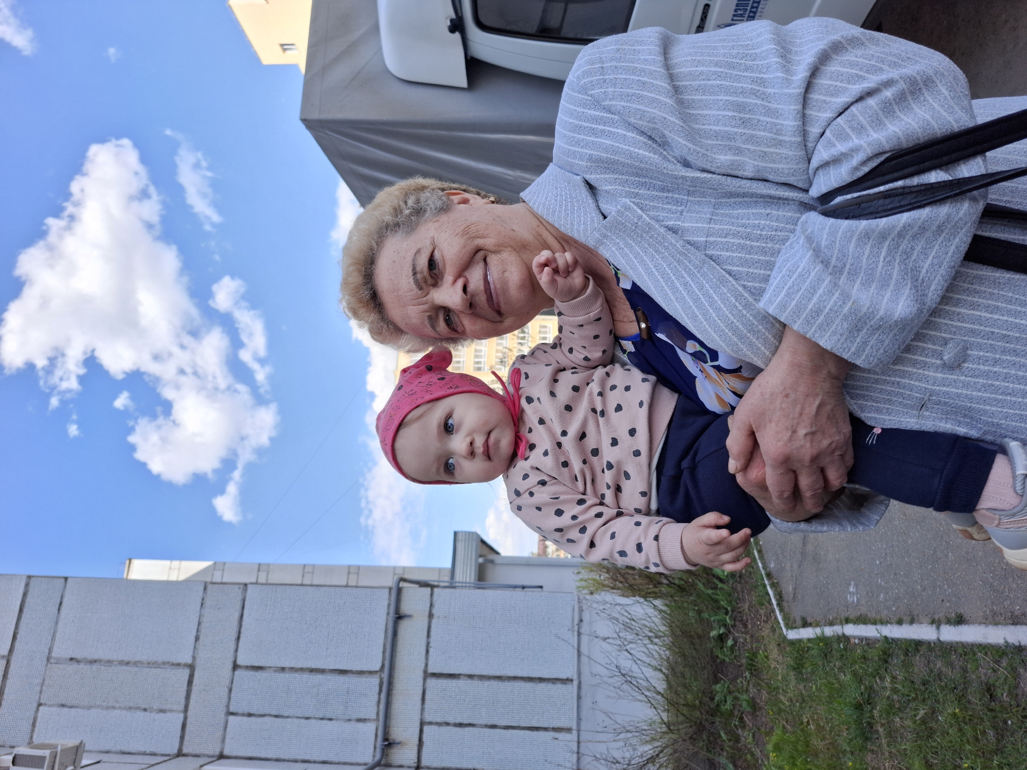 An elderly woman with short blonde hair, wearing a grey pinstripe blazer, holds a baby in her arms. The baby is wearing a pink hat and a pink sweater with black polka dots, and dark blue pants. The background shows a partially visible white truck and a building with white rectangular blocks, a blue sky with white clouds, and some greenery. The location is determined to be Chaikovsky, Russia based on markings on the visible part of the truck.