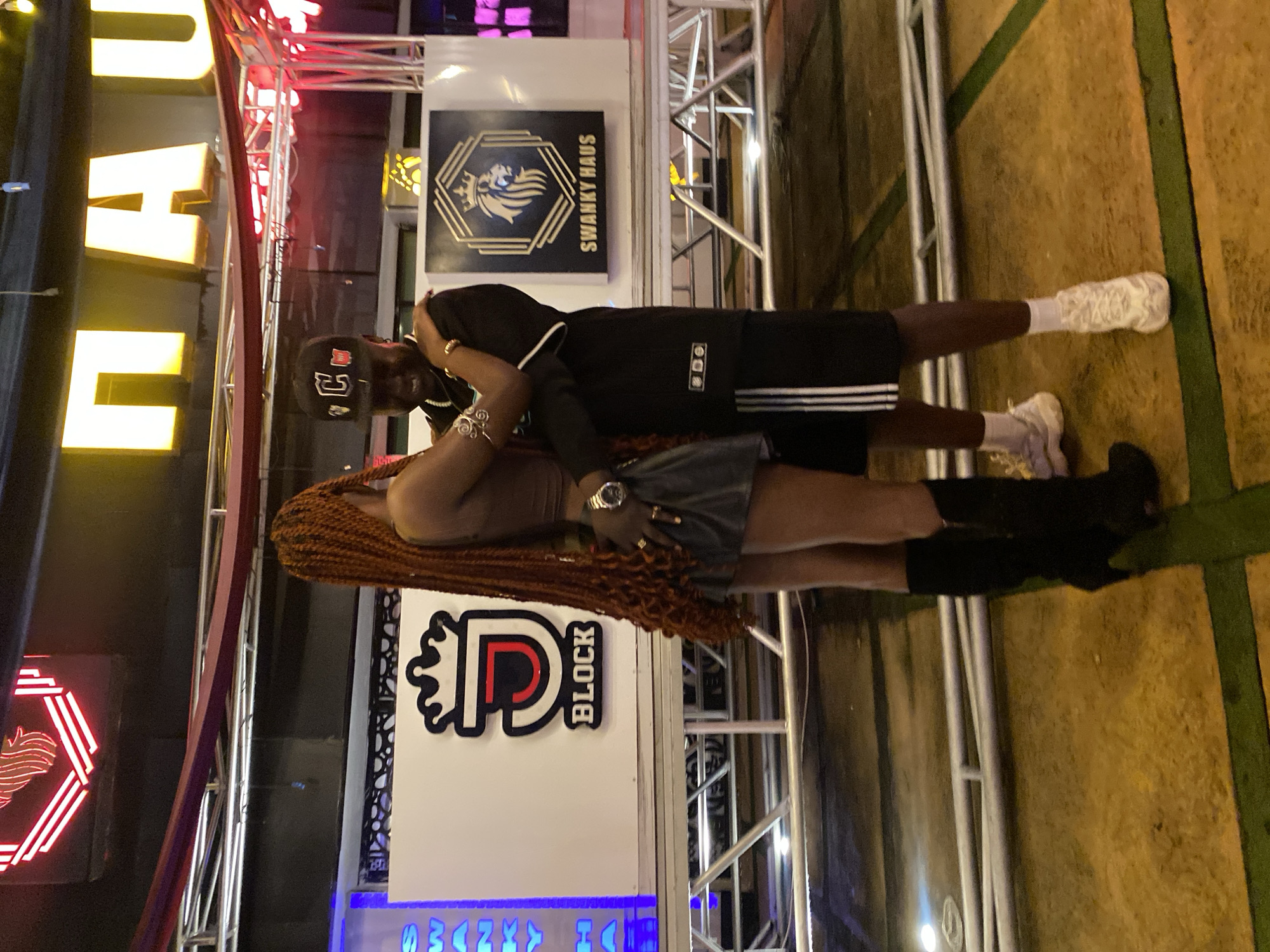 In Bar Jirgi Summa, Nigeria, two individuals, a woman and a man, are embracing. The woman, with long, dark braids, wears a brown top and a dark skirt. The man, wearing a black baseball cap, a black long-sleeved shirt with white stripes on the sleeves, and black shorts with white stripes, has his arm around the woman. They are positioned in front of a white backdrop with two logos. The left logo features a stylized "J" with a crown above it and the text "J BLOCK" below. The right logo is a hexagonal emblem with a lion and the text "SWANKY HAUS." Behind them, a large neon sign reads "NAU" in yellow block letters against a dark background, with red neon accents above it. The scene appears to be an outdoor venue, possibly a club or entertainment space, with metal scaffolding and lighting structures visible.