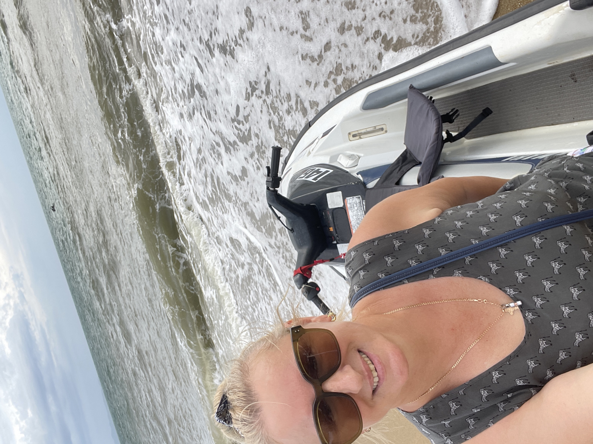 A blonde woman wearing sunglasses and a dark grey sleeveless dress with a zebra pattern smiles at the camera while sitting on a jet ski on a beach. The jet ski is white and grey with black handlebars and a grey seat. The ocean is in the background with waves crashing on the shore. The sky is overcast. The location is Pattaya City, Thailand.
