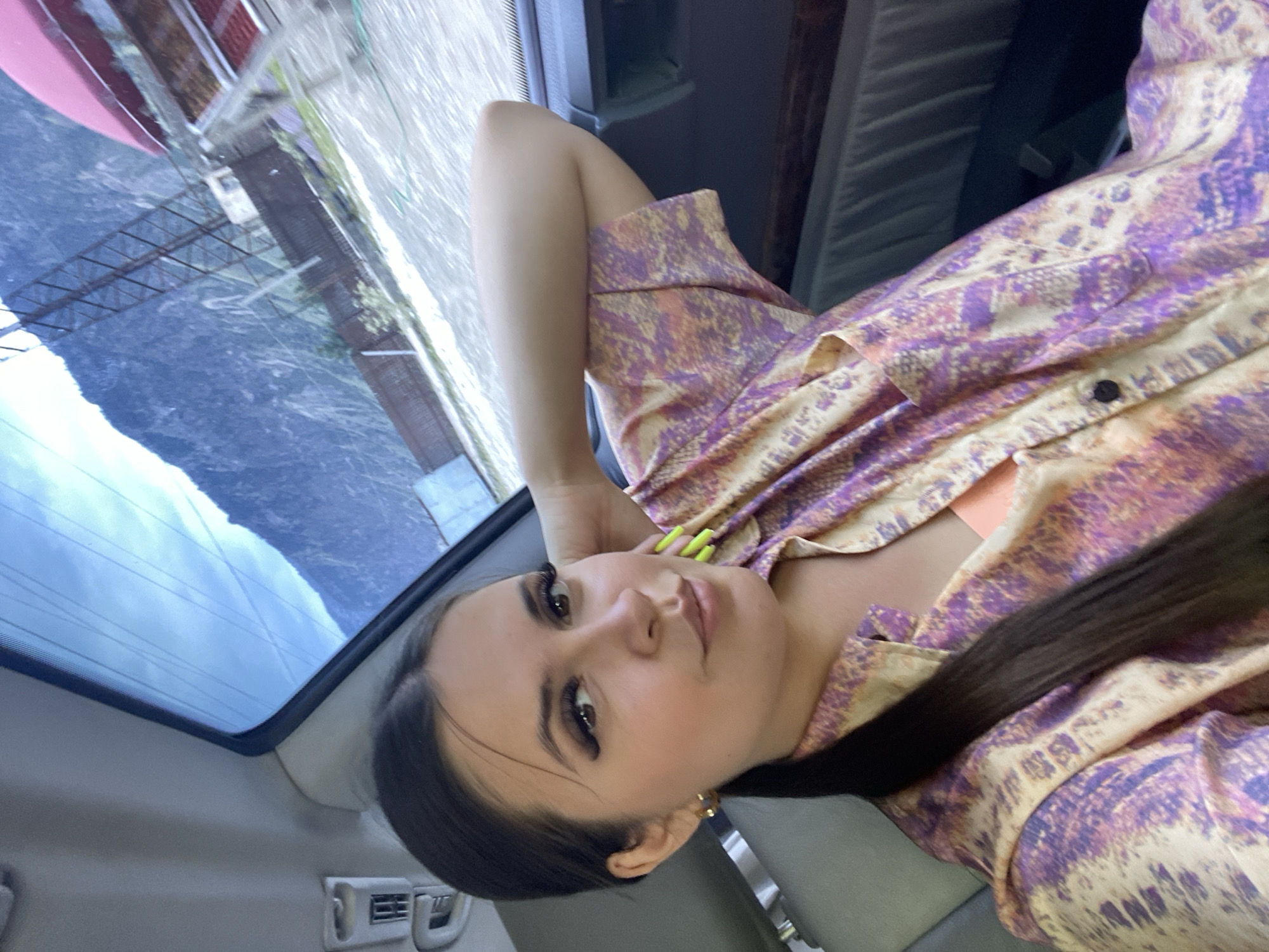A young woman with dark hair pulled back in a ponytail is seated in the passenger seat of a vehicle, her right arm resting behind her head. She is wearing a patterned shirt with shades of purple, orange, and cream, and an orange camisole is visible underneath. Her nails are painted a neon yellow-green. The interior of the car is dimly lit, with a glimpse of a grey leather seat. Through the rear window, a mountainous landscape under a cloudy sky is visible. The exterior scene shows a dirt or gravel road, a fence, some structures, and a tall metal pylon. The overall impression is a casual, candid shot taken during a road trip in a scenic, mountainous region. The location is identified as Irganai, Russia.