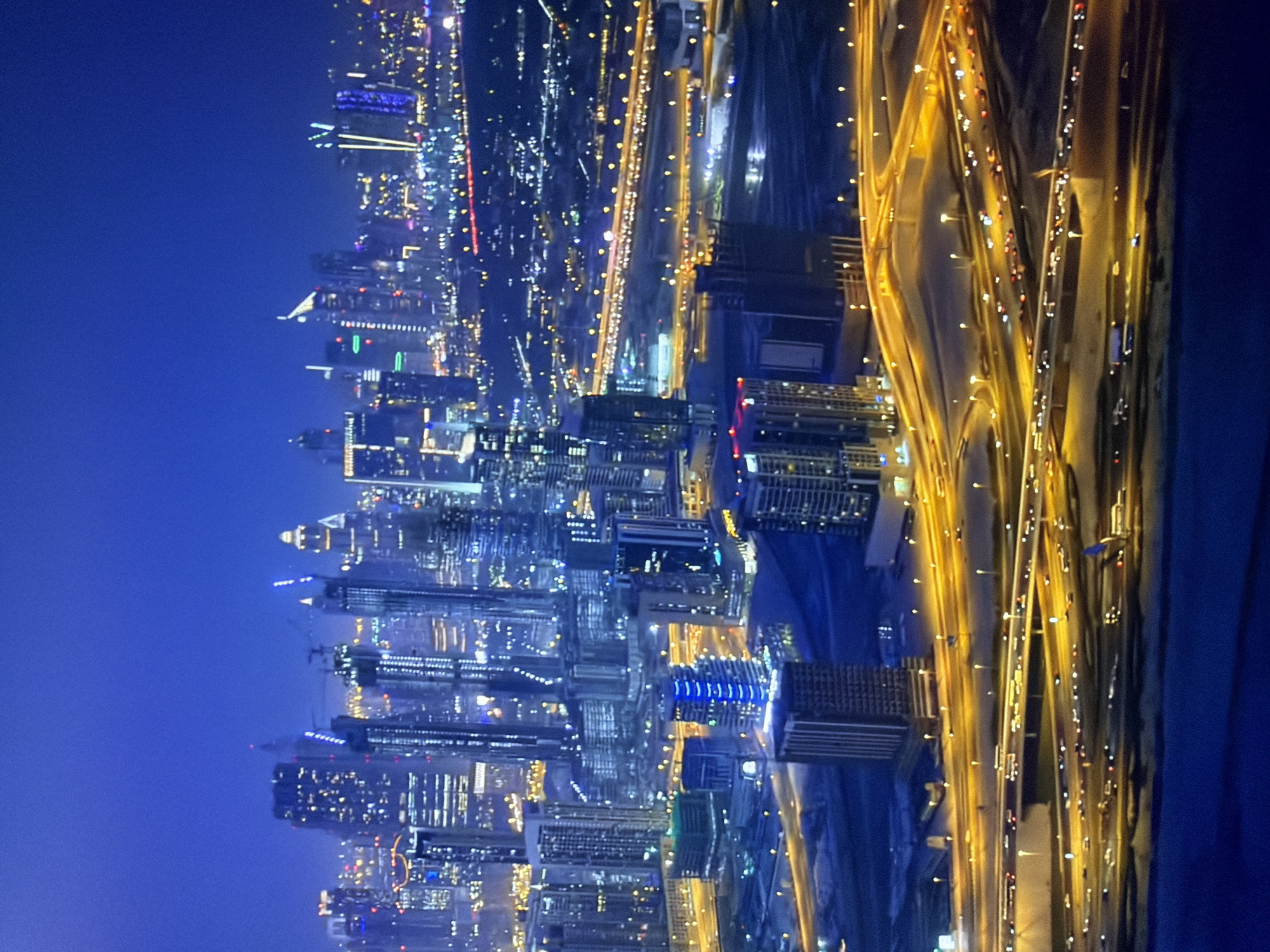 This is a nighttime aerial view of a modern city skyline at dusk or night. The sky is a deep, saturated blue, fading to a lighter hue near the horizon, suggesting twilight or a clear night. A dense cluster of tall skyscrapers dominates the scene, their windows illuminated with a mix of white, yellow, and blue lights, creating a vibrant, glowing effect. Some buildings appear to be under construction, with visible cranes.
Below the skyscrapers, a complex network of multi-level highways is illuminated by bright yellow streetlights and the headlights of numerous cars. The roads curve and intersect, creating a dynamic visual pattern. The ground surrounding the highways appears to be a mix of dark land and possibly water or water features, reflecting some of the city lights. The overall impression is one of urban development and activity, with the city lights creating a striking contrast against the dark night sky. The image does not depict the city of Kyiv, Ukraine, but rather a city with a very modern and advanced urban landscape, possibly in the Middle East or Asia, given the style of architecture and the density of the development.