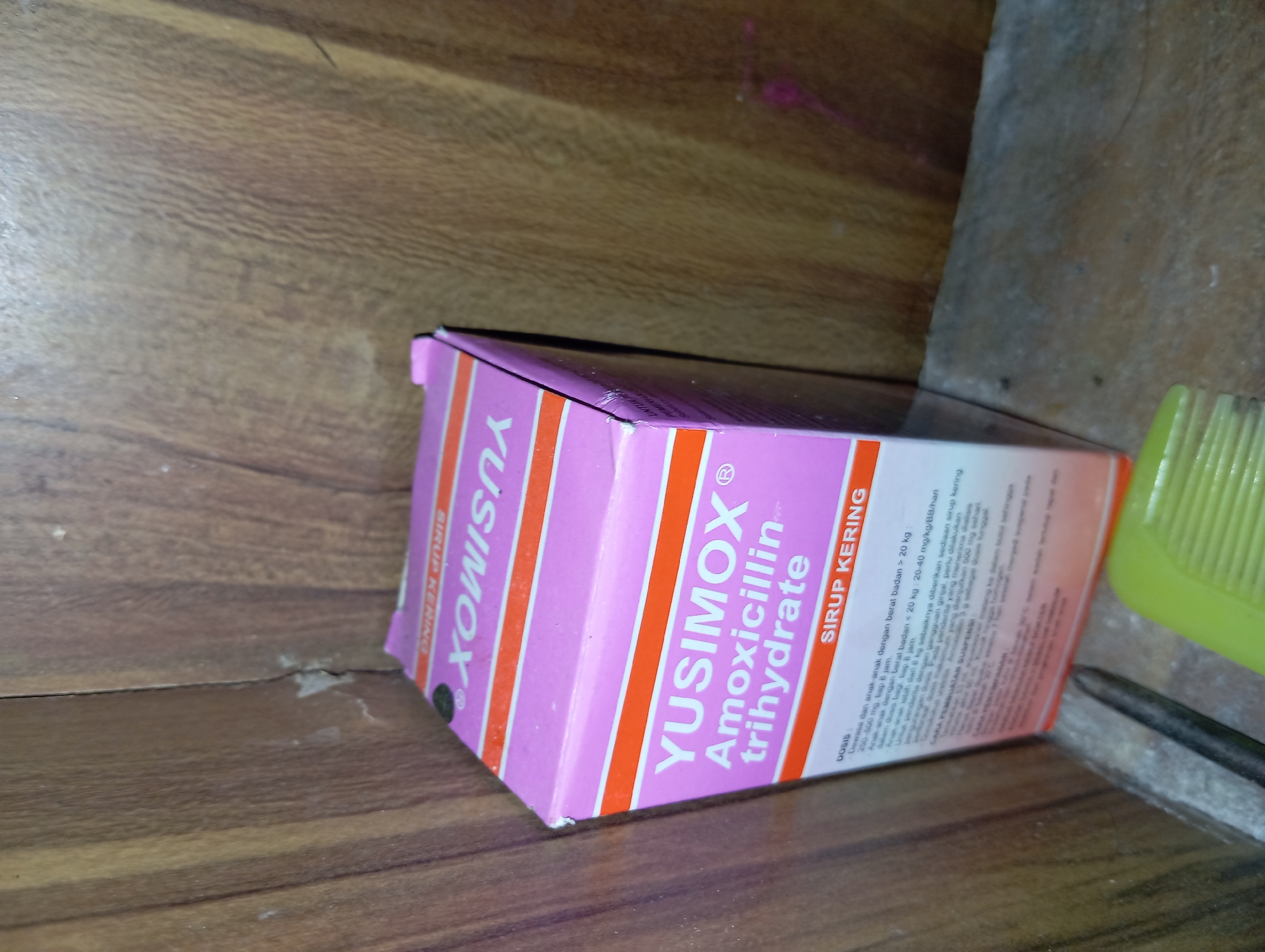 A pink and orange box for YUSIMOX Amoxicillin trihydrate SIRUP KERING is placed against a wooden textured background. The box is slightly tilted. Some text is visible on the box, including dosage instructions. A green plastic object, possibly a comb or brush, is visible in the bottom center of the image, partially obscuring the box. The background appears to be a wall or shelf with wood grain texture. There is a slight shadow on the left side of the image, suggesting lighting from the right. The image is taken from a slightly elevated angle, looking down at the box. The scene appears to be indoors, possibly a home environment. There are no people or animals visible in the image. The lighting is moderate, with some shadows. The overall impression is one of domestic clutter, with the medication box being the central focus. It is not possible to determine the exact location in Rejosari, Indonesia, based solely on the visual information provided.