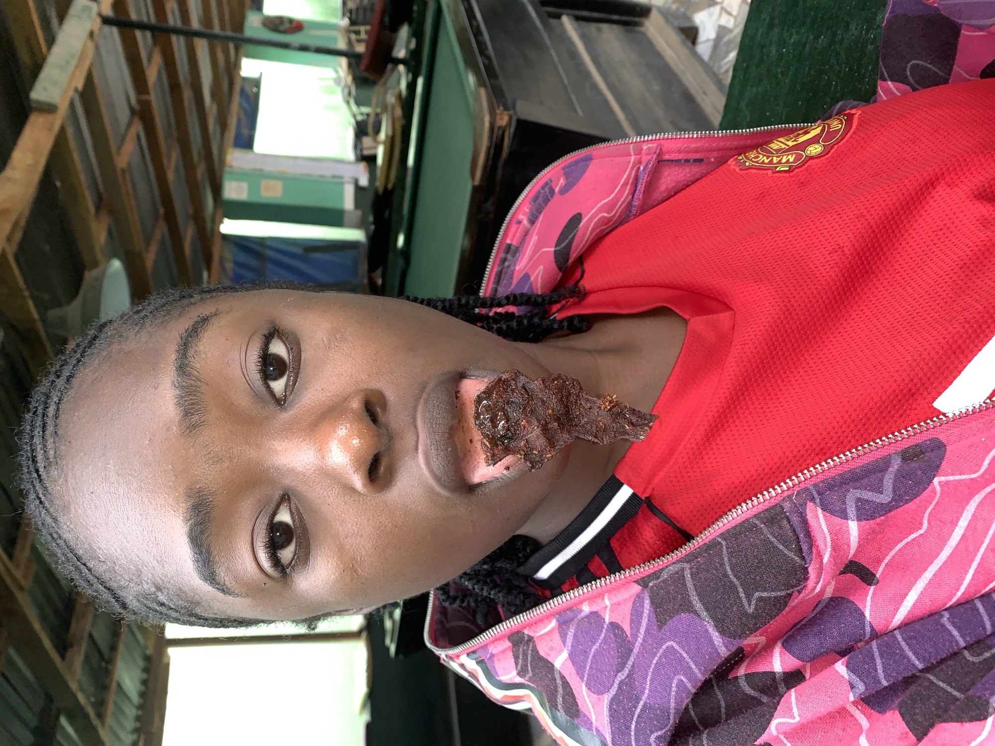 The image displays a young individual, likely female, with dark skin and braided hair, posing for a photograph in an indoor setting. The image is rotated 90 degrees counter-clockwise. A piece of dark, possibly dried or cooked, meat is visible on their tongue or extending from their mouth, creating a casual or playful pose.
The person is wearing a red short-sleeved top, which appears to be a football jersey, featuring a "MANCHESTER UNITED" logo on the left chest, characterized by a golden lion and football emblem. Over this jersey, they wear an open, zipped jacket or hoodie with a vibrant and distinct pattern of pink, purple, black, and white abstract shapes. The individual looks directly at the camera with a neutral expression.
The background shows wooden structures, possibly shelves or partitions, suggesting a retail or storage area. A bright light source or opening is visible behind them, indicating it is daytime. The overall setting is consistent with an informal indoor environment, likely in Jos, Nigeria.