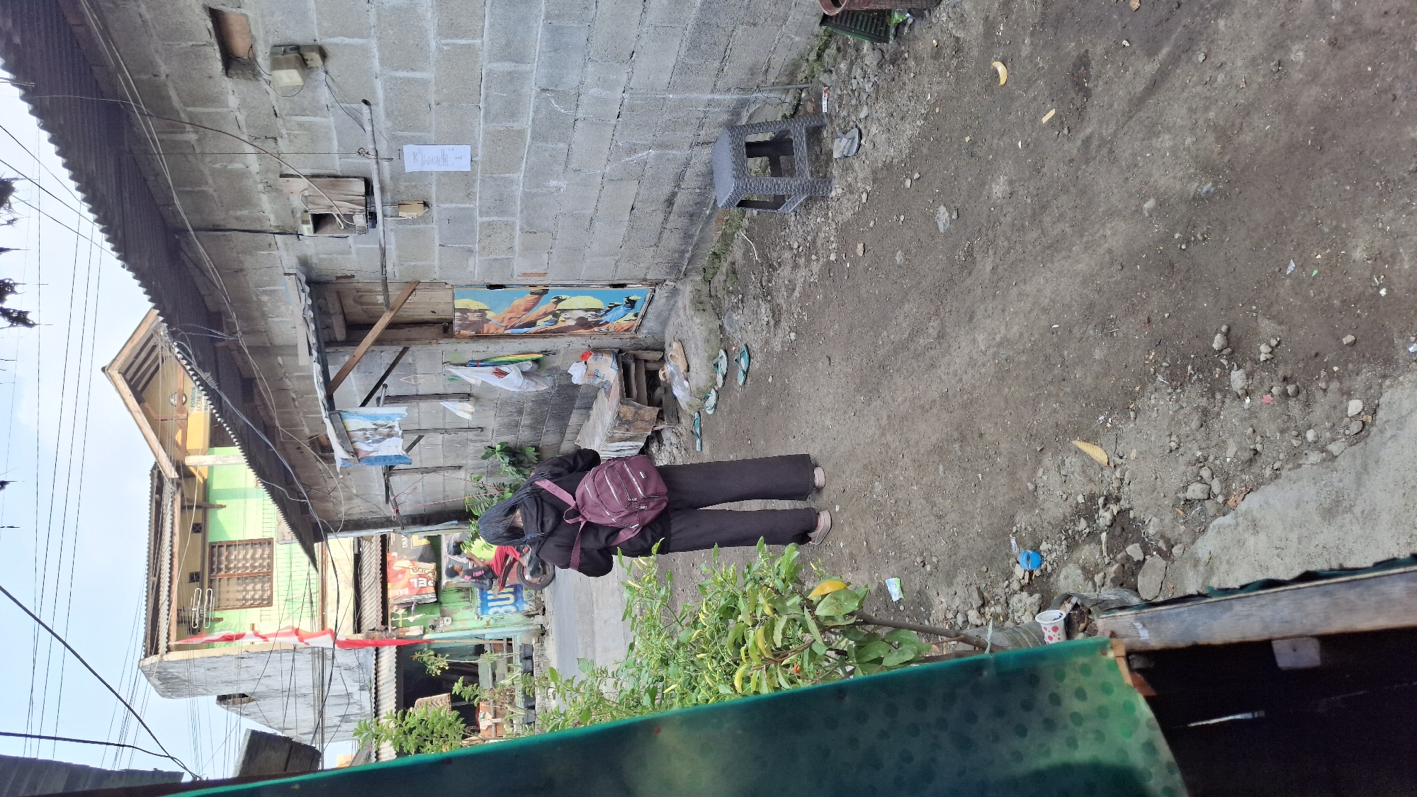 The image depicts an outdoor scene on a dirt path in Wadungasri, Indonesia. A person wearing a dark hoodie and loose black pants is standing with their back to the camera, carrying a maroon backpack. They are looking towards a street where a red motorcycle with a person wearing a black shirt and pink pants is visible. To the right of the path, a wall constructed of cinder blocks forms the side of a building. Several items are placed against this wall, including a small plastic stool, a wooden bench, and various pieces of clothing or fabric hanging from a line. A colorful mural or poster is also attached to the wall. Scattered on the ground are several pairs of flip-flops, some scattered rocks, and debris. The environment suggests a residential area with simple construction. The time of day appears to be late afternoon or early evening, given the lighting.