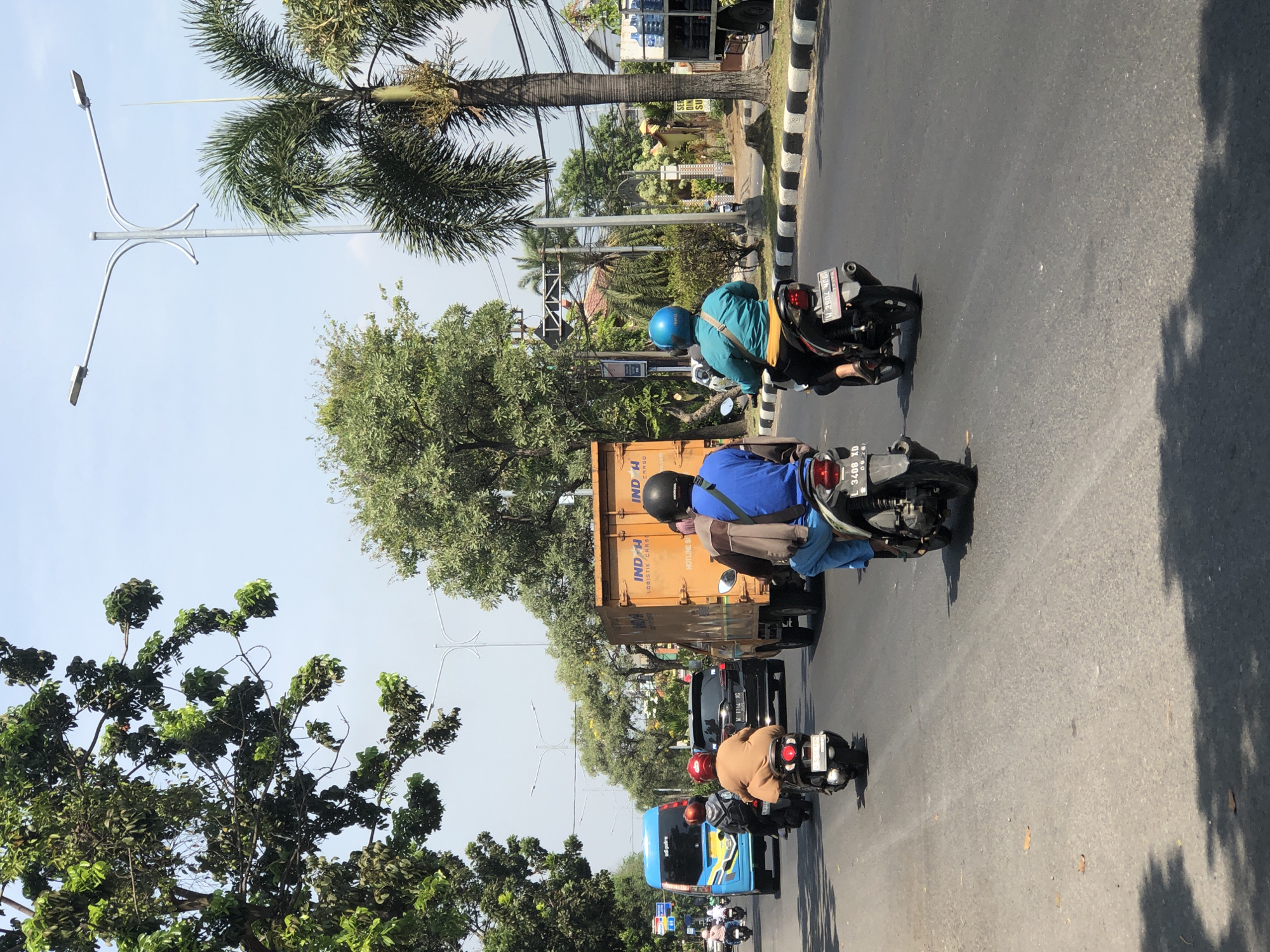 This image depicts a street scene in Larangan, Indonesia, on a bright, sunny day. The perspective is from behind moving vehicles on a paved road. In the foreground, two motorcycles are visible. The motorcycle on the left has a license plate that reads "3400 XD". The motorcycle on the right, with a rider wearing a blue helmet and teal shirt, has a license plate "V 2604 JC". Further ahead, a large orange truck with "INDH LOGISTIK CARGO" written on its side is driving. To the left of the truck, a blue bus with "edi putra" on its side is also in motion. Several trees line the right side of the road, including palm trees, and a street lamp with two lights is prominent. The sky is clear and blue. The road is marked with white and black striped curbs. The overall impression is one of moderate traffic in an urban or suburban environment.