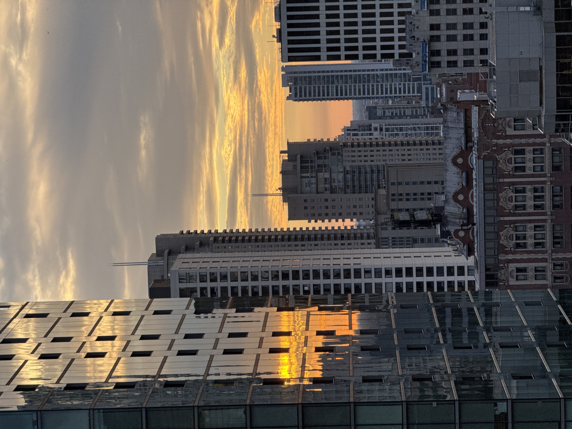 This is a photograph of a city skyline, likely Chicago, United States of America, during sunset. Tall buildings of varying architectural styles dominate the frame. On the left, a modern skyscraper with a glass facade reflects the warm orange and yellow hues of the setting sun, creating a striking visual effect. To its right, several other high-rise buildings are visible, including some with white exteriors and numerous windows, and one with a black and white striped facade. In the background, the sky is filled with clouds, painted in shades of orange, yellow, and gray, indicating the sun is low on the horizon. The overall scene captures a city at dusk, with the urban landscape illuminated by the fading light.
