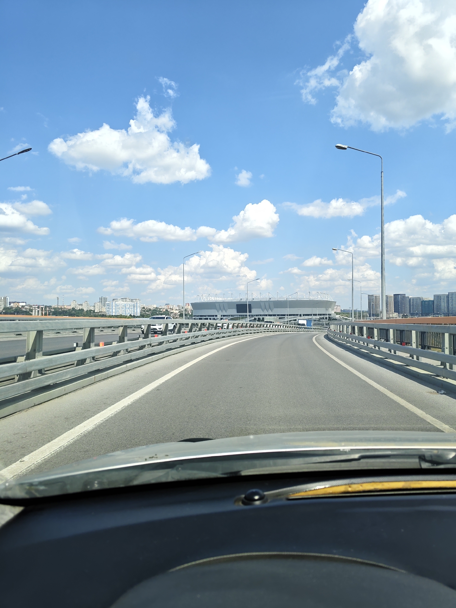 The image captures a view from a vehicle driving on a modern overpass, likely a bridge, on a clear day with a bright blue sky dotted with fluffy white clouds. The overpass features a paved road with white lane markings and a metal guardrail running along the right side. Streetlights are visible at intervals along the overpass. In the distance, a large, modern stadium with a distinctive roof structure is prominent. Surrounding the stadium are numerous tall, modern buildings, suggesting an urban setting. The foreground of the image shows the dashboard and windshield of the vehicle, partially obscuring the view. The architecture of the buildings and the design of the stadium suggest it might be the Rostov Arena, a stadium built for the 2018 FIFA World Cup, located in Rostov-on-Don, Russia.