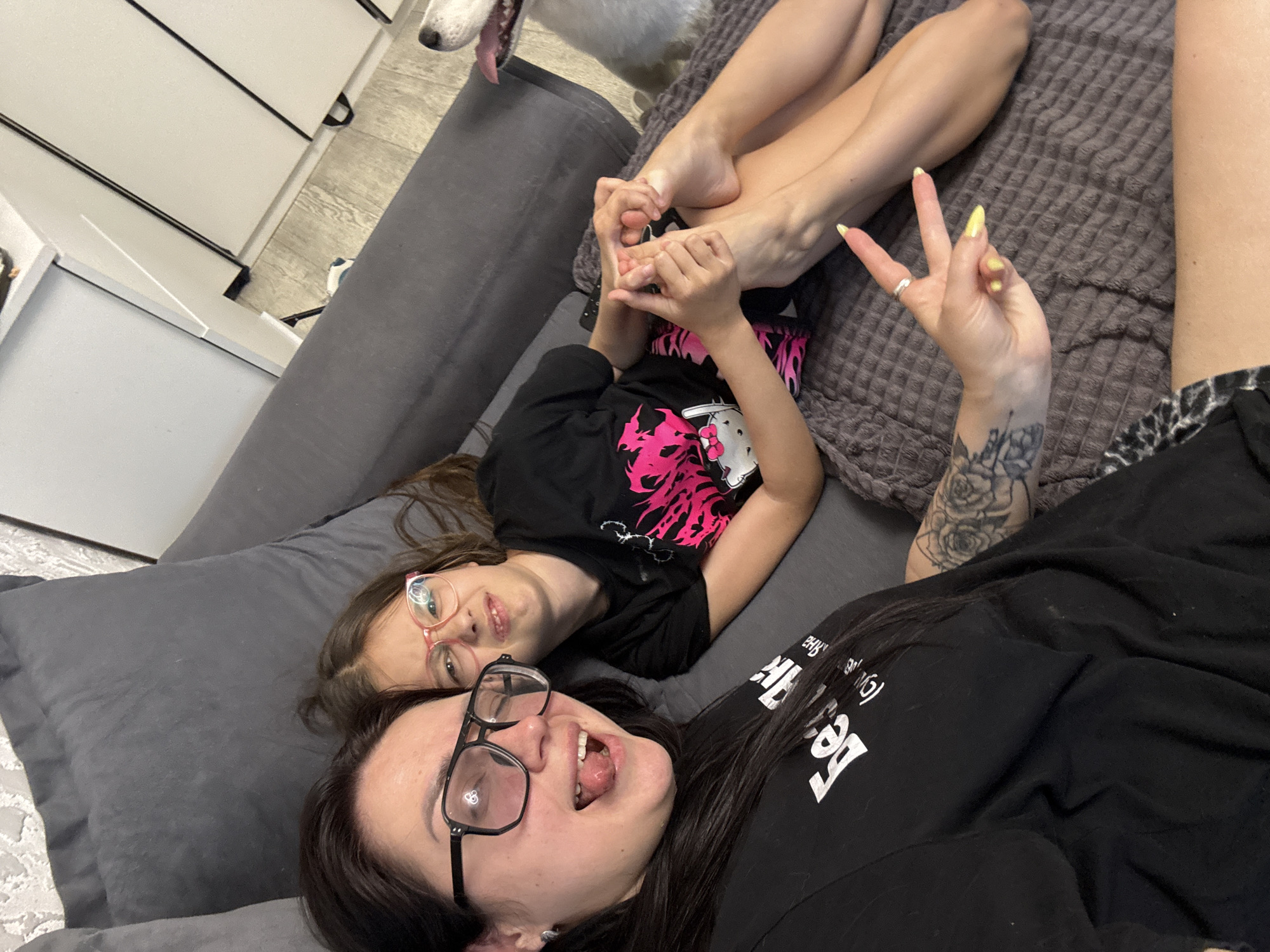 The image captures a moment of leisure indoors, likely a residence in the city of Unknown, Unknown, as no specific location indicators are present. A woman with dark hair and glasses is seen in the foreground, lying on a grey upholstered surface that appears to be a couch or bed. She is wearing a black t-shirt with white text and has a tattoo on her upper arm. Her mouth is open, and she is making a playful expression. Next to her is a young girl, also lying down, wearing glasses and a black t-shirt with a pink and white design. Their legs are intertwined. The woman has her hand up, making a gesture. In the background, there's a glimpse of a white dog with its mouth open, suggesting it might be panting or barking. The environment consists of a grey quilted blanket or padding and a partially visible white cabinet with black handles and trim in the background. The overall lighting appears to be bright, likely from indoor sources.