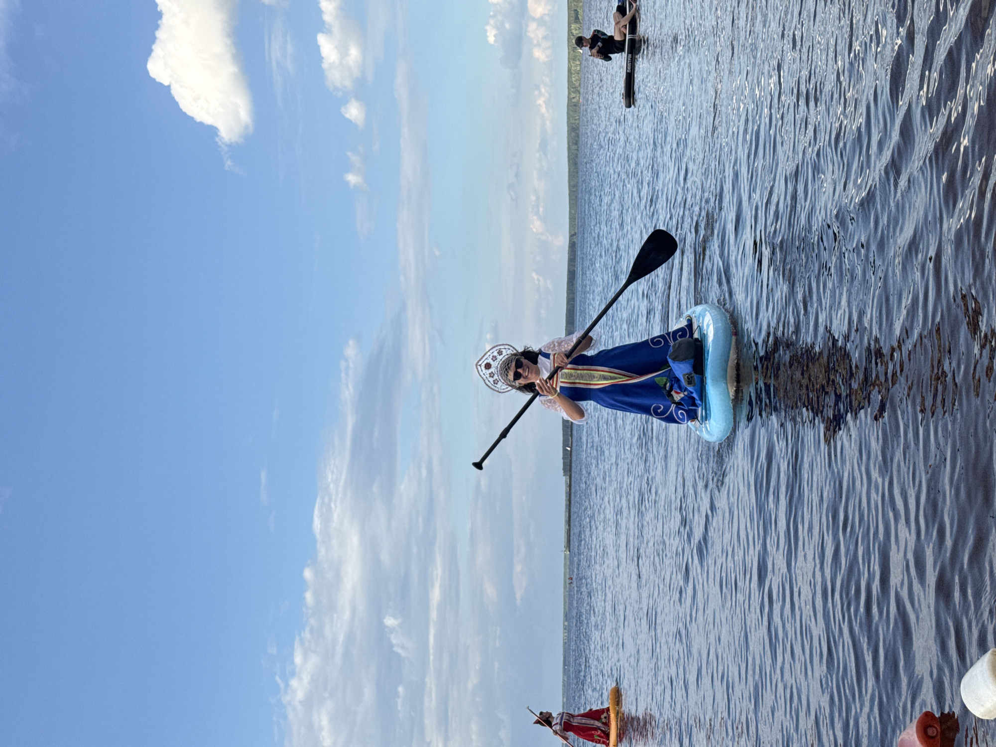 In Chaikovsky, Russia, on a sunny day with a partly cloudy sky, several people are paddleboarding on a wide, calm body of water, likely the Kama River. In the foreground, a woman dressed in a blue traditional Russian sarafan and a decorative kokoshnik headdress stands on a paddleboard, holding a paddle. She is looking at her phone, which she is holding up. Another paddleboarder, also in traditional attire, is visible in the middle ground on the left, wearing a red dress and holding a paddle. In the background on the right, a person in dark athletic wear is sitting on a paddleboard. The water is a deep blue with gentle ripples. Several buoys or flotation devices are visible in the lower left corner of the image. The horizon line is flat, indicating a large expanse of water and a distant treeline.