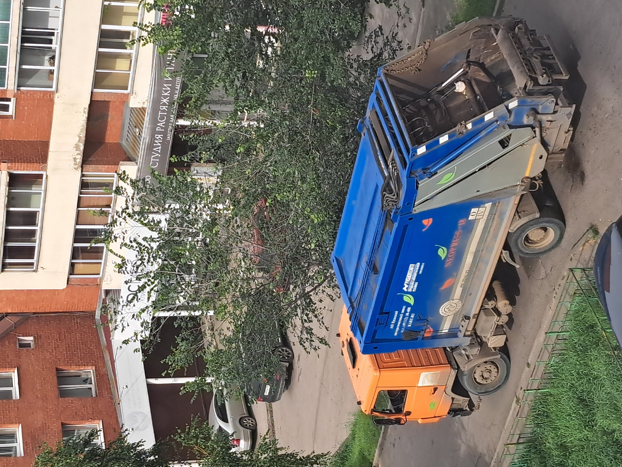 A garbage truck with an orange cab and a blue compactor body is parked on a street in front of a brick apartment building. The truck has Cyrillic lettering on its side, indicating it is from AO Ryazan. The street is paved, and there is a patch of green grass and a metal fence to the left of the truck. In the background, trees with green foliage partially obscure the building. A silver sedan and a dark SUV are parked on the street behind the garbage truck. The apartment building has multiple stories with windows, and a sign with Cyrillic lettering is visible above the entrance. The scene appears to be a typical urban environment in Irkutsk, Russia, during daylight hours.