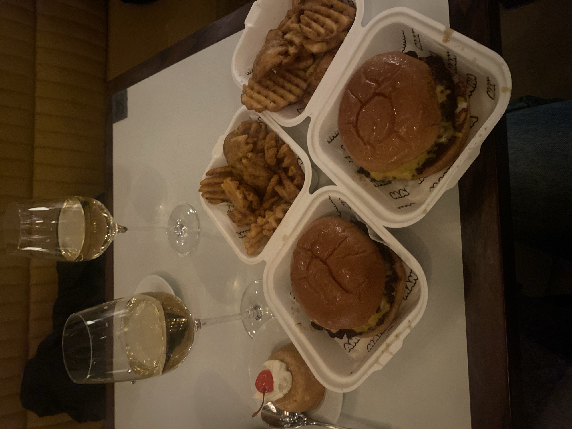 The image shows a table setting with food and beverages. There are two glasses of white wine. There are two burgers, two portions of waffle fries, and a small dessert with a cherry on top, all served in white takeaway containers. The table is white and bordered by a dark wood trim. The photo was taken indoors. The restaurant appears to be in London, United Kingdom.