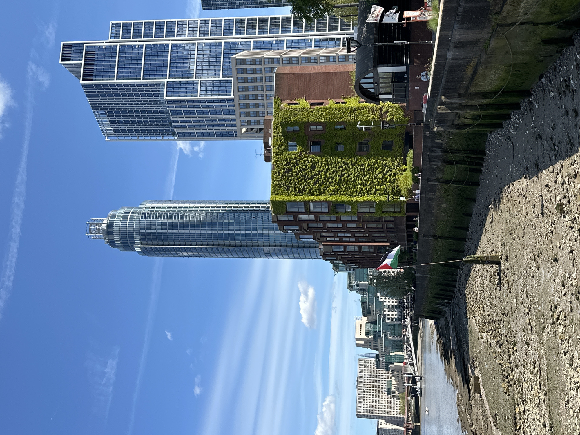 The image shows a cityscape along a riverbank in London, United Kingdom. The camera appears to be positioned along the riverbank, capturing a view of modern buildings.
On the left side of the image is a clear blue sky. Dominating the center are several skyscrapers of varying heights and designs. One building is partially covered in lush green foliage. A flag is visible on the riverbank in the middle ground.
On the right, the image includes a textured embankment or river wall, suggesting low tide conditions and revealing a rocky or pebbly surface.
A river is visible, likely the Thames, with some boats visible in the background. In the background, more buildings form the cityscape.