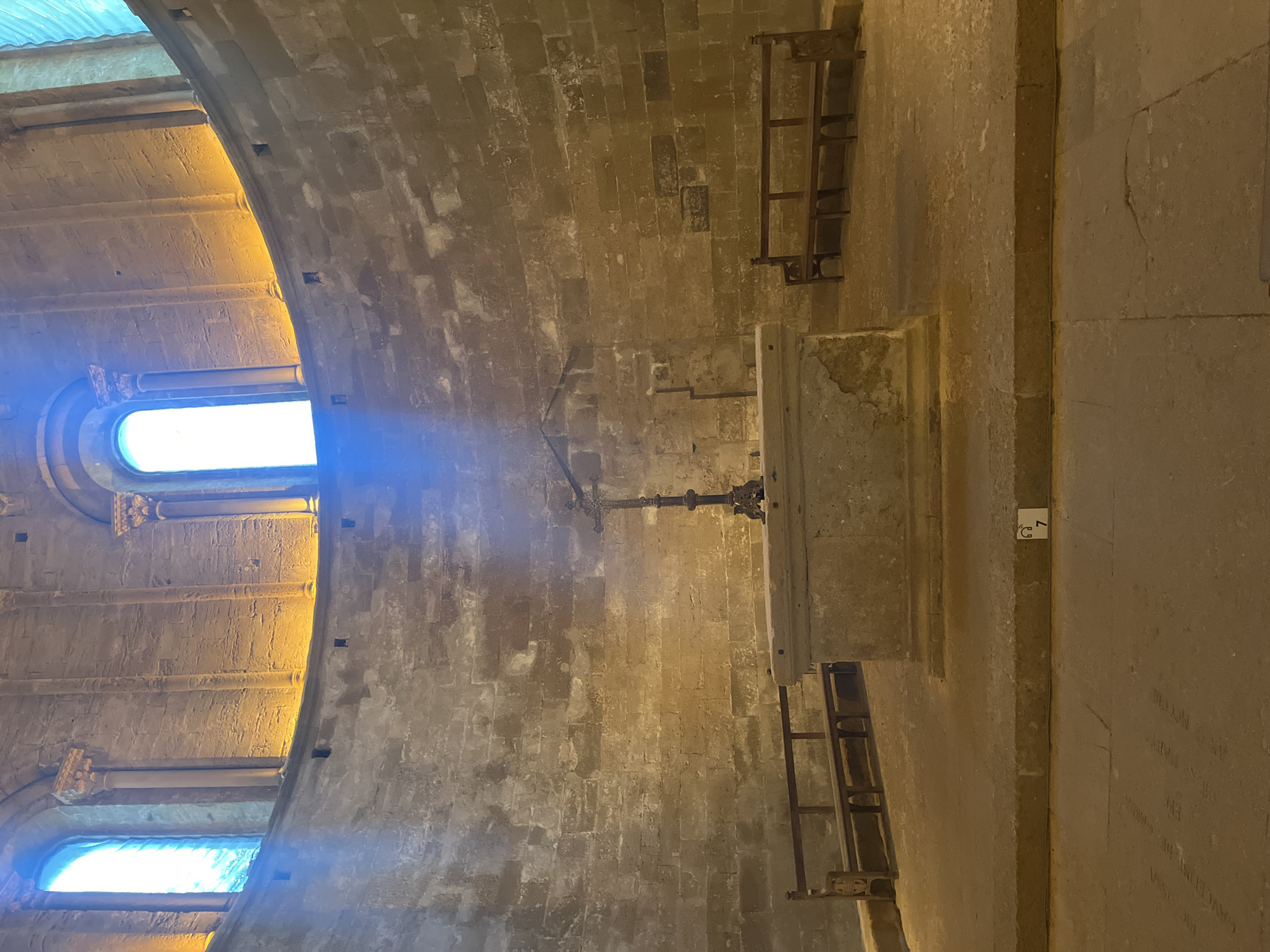 The image shows the interior of a circular stone chamber, likely part of a historical religious building. Two tall, arched windows with blue-tinted glass are visible at the top of the curved wall, with warm yellow light illuminating the upper portions of the stone. In the center of the chamber is a simple stone altar or pedestal. A dark, ornate metal crucifix stands on the altar. Two wooden benches with simple backrests are positioned against the curved walls, one on the left and one on the right. The walls are constructed of rough-hewn stone blocks. The floor is also made of large stone slabs. A small sign with a headphone icon and the number "7" is visible on the raised platform in front of the altar. The overall impression is one of age and solemnity, with a focus on the architectural elements and the religious artifacts. This scene is likely located within the ruins of the Saint-Pierre de Maguelone Cathedral in Villeneuve-lès-Maguelone, France, a site known for its Romanesque architecture and historical significance.