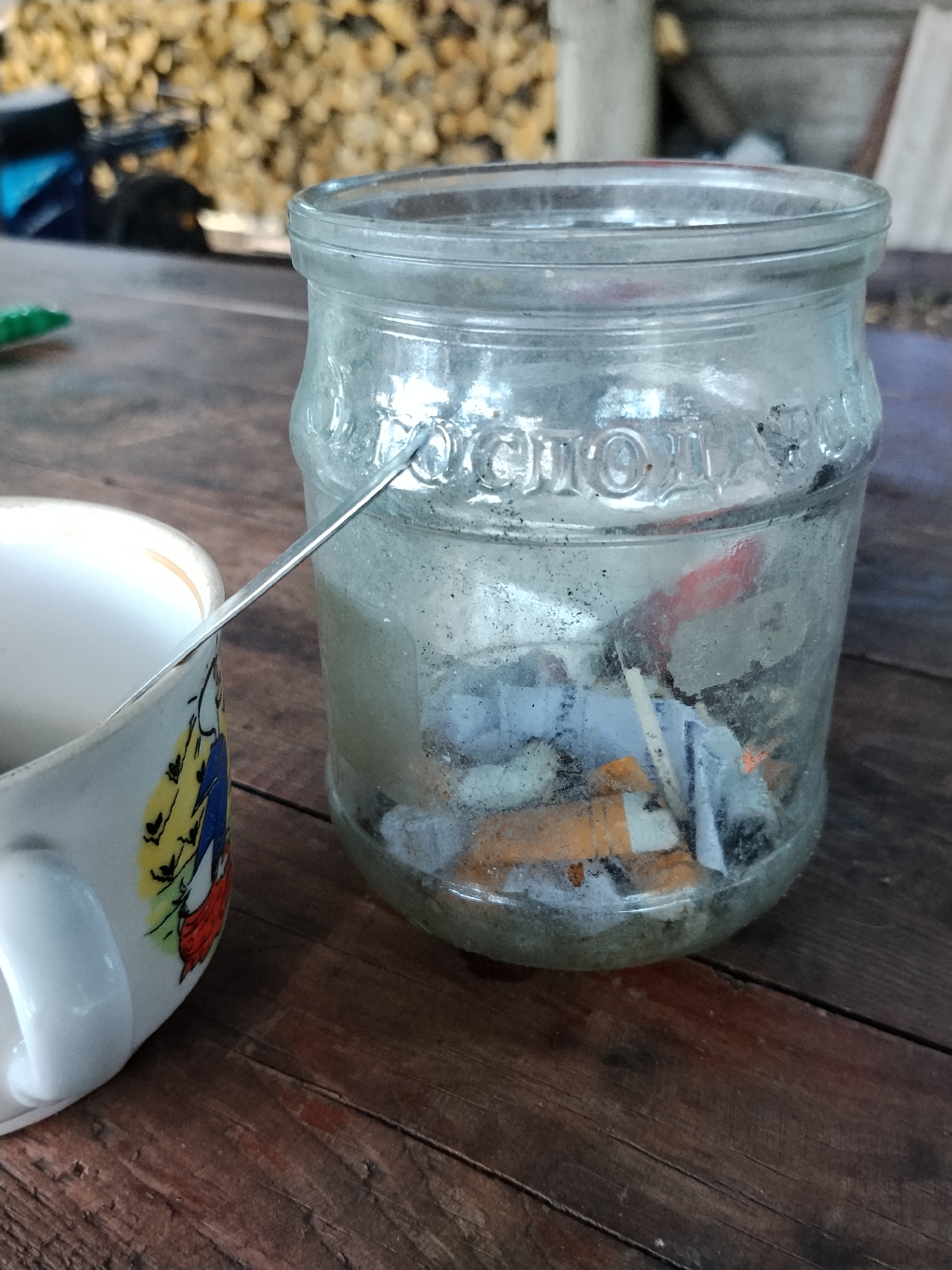 An outdoor, close-up shot shows a glass jar filled with cigarette butts and water, positioned on a weathered wooden table. A metal spoon extends from the jar towards a white ceramic mug, which is decorated with a colorful, abstract design featuring yellow, blue, and red elements, and a white handle. The background is blurred, showing a pile of firewood and indistinct structures, suggestive of a rural or semi-urban setting. The scene appears to be located in or around the city of Hoholeve, Ukraine.