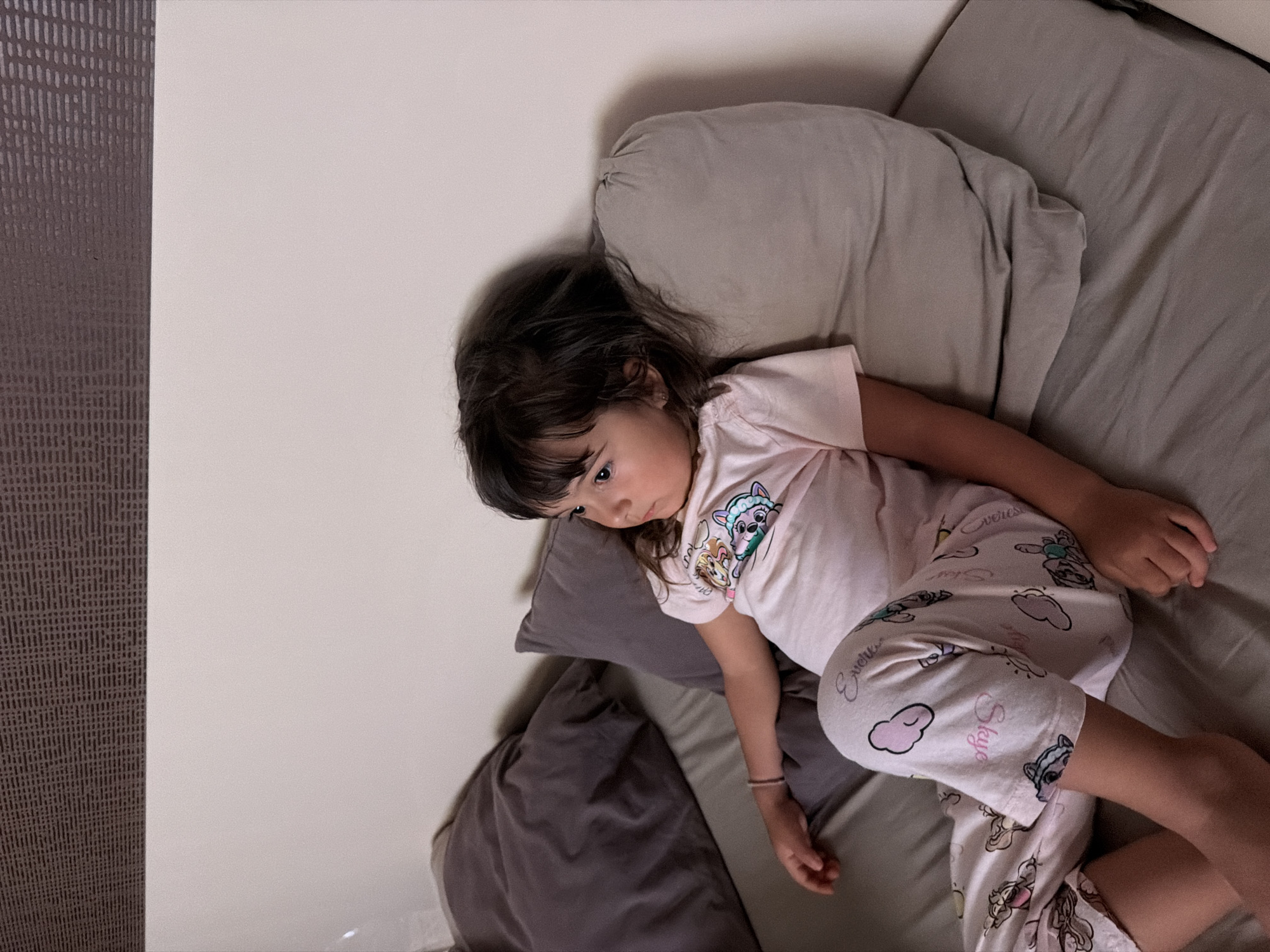 The image shows a young girl lying on a bed, facing right. She is wearing pink pajamas with cartoon characters and the word "Skye" printed on them. The bed is made with grey sheets and has several pillows. One pillow is behind the girl's back, another is to her right, and a larger, darker pillow is to her left. The background consists of a plain white wall and a textured brown headboard. The lighting suggests it is either nighttime or indoors with dim lighting. The girl's hair is dark brown and falls over her shoulders. She has a small pink bracelet on her left wrist. The scene appears to be a bedroom. No specific location in Burgas, Bulgaria, can be identified from the image.