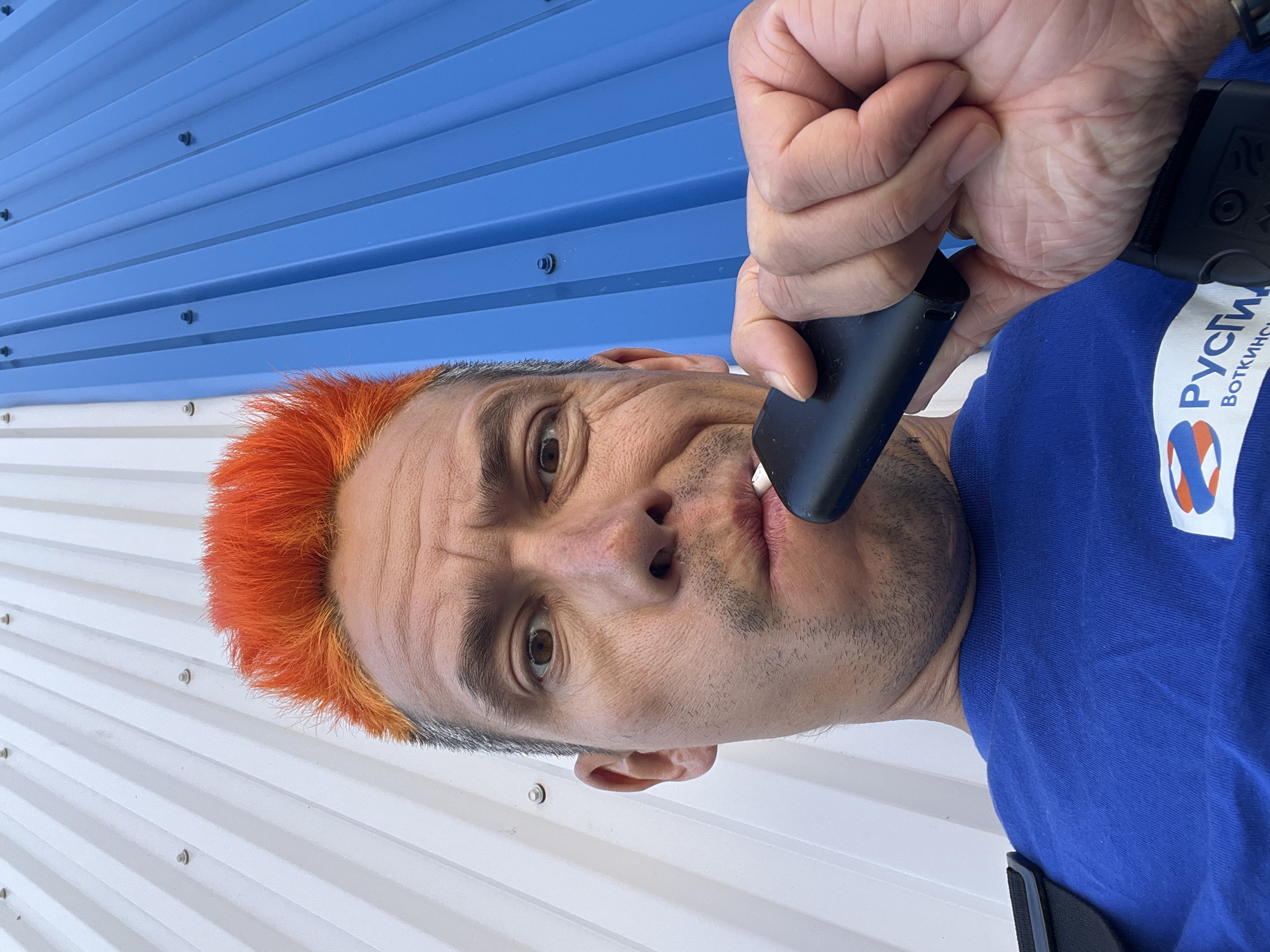 A man with orange hair is shown in a close-up selfie. He is wearing a blue t-shirt with a logo on it. He is holding a black object, possibly a vape pen, to his lips. The background is a blue and white corrugated metal wall. The logo on the shirt is partially obscured but appears to say "Рустик." The location in Chaikovsky, Russia cannot be determined from the image.