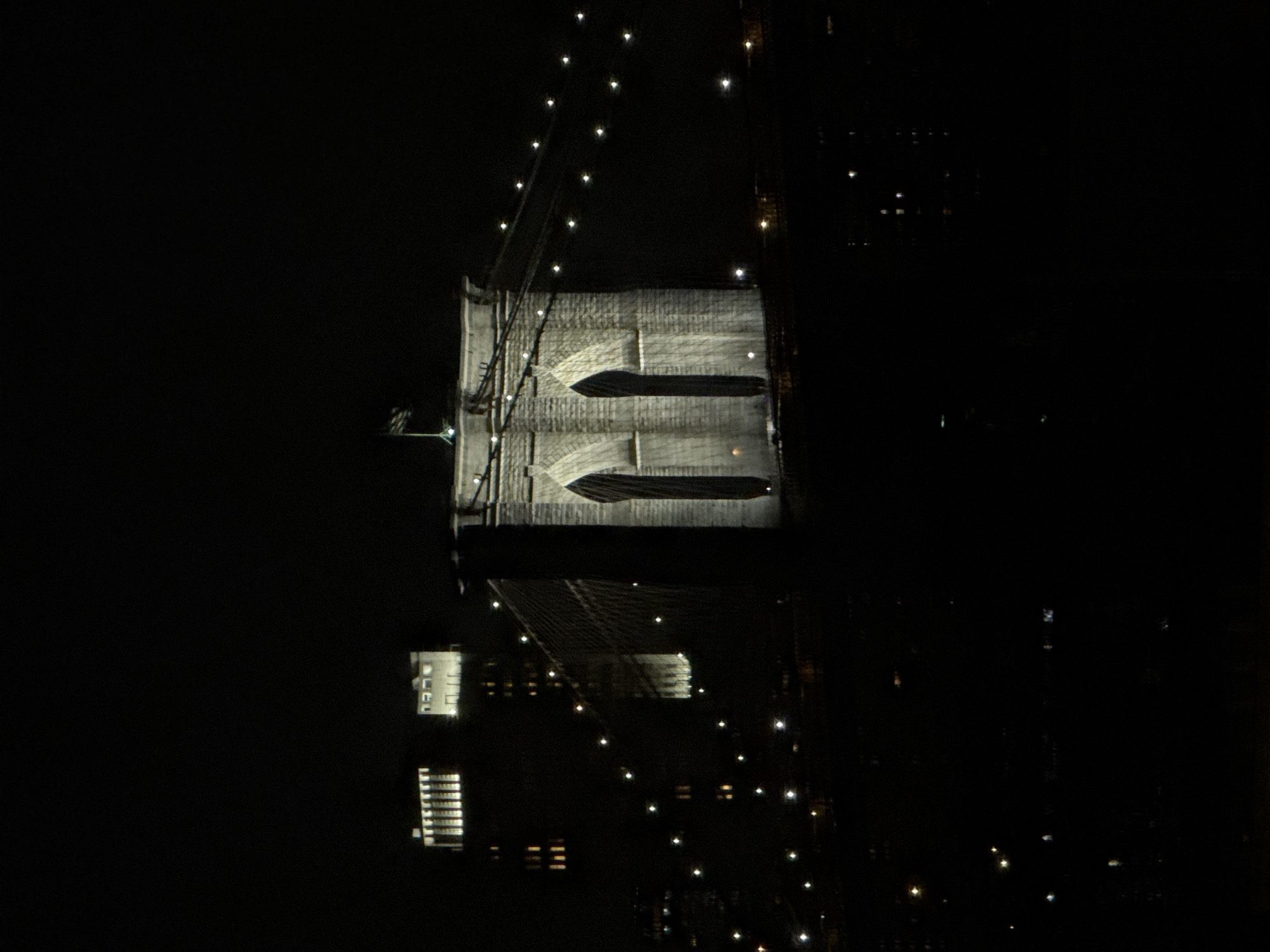 Here is a description of the image:
This image depicts a nighttime view of the Brooklyn Bridge in New York City. The image is mostly dark, with the bridge illuminated by lights along the cables and towers. Parts of the surrounding cityscape are also visible, with lights from buildings scattered in the background. The architecture of the bridge is prominently featured, showing its support structure and design. The image appears to be taken from an aerial perspective.