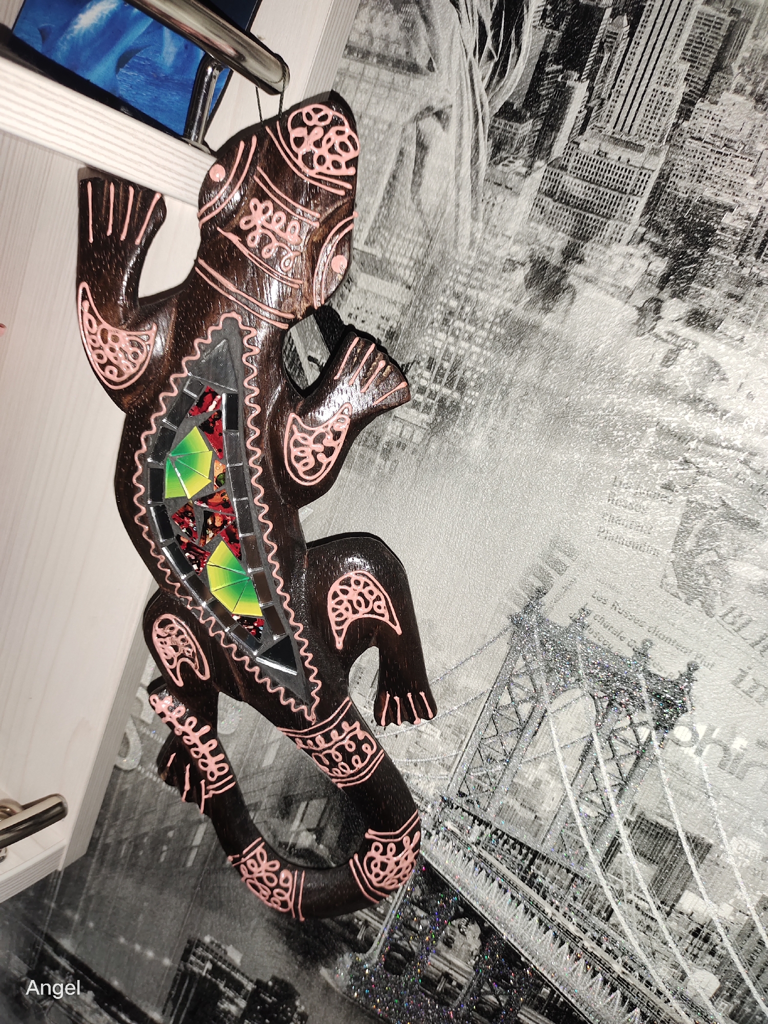 A dark wood, mosaic gecko-shaped ornament hangs from a metal rod, its body adorned with pink decorative patterns and a mosaic inlay of colorful glass pieces. The ornament is positioned against a black and white mural depicting a cityscape and a bridge, with a silvery glitter overlay. To the left, a white shelf with a silver handle is visible, and a blue and black image is partially seen. The bottom left corner of the image has the word "Angel" in white text. The scene is indoors, and there is no indication of Tula, Russia.