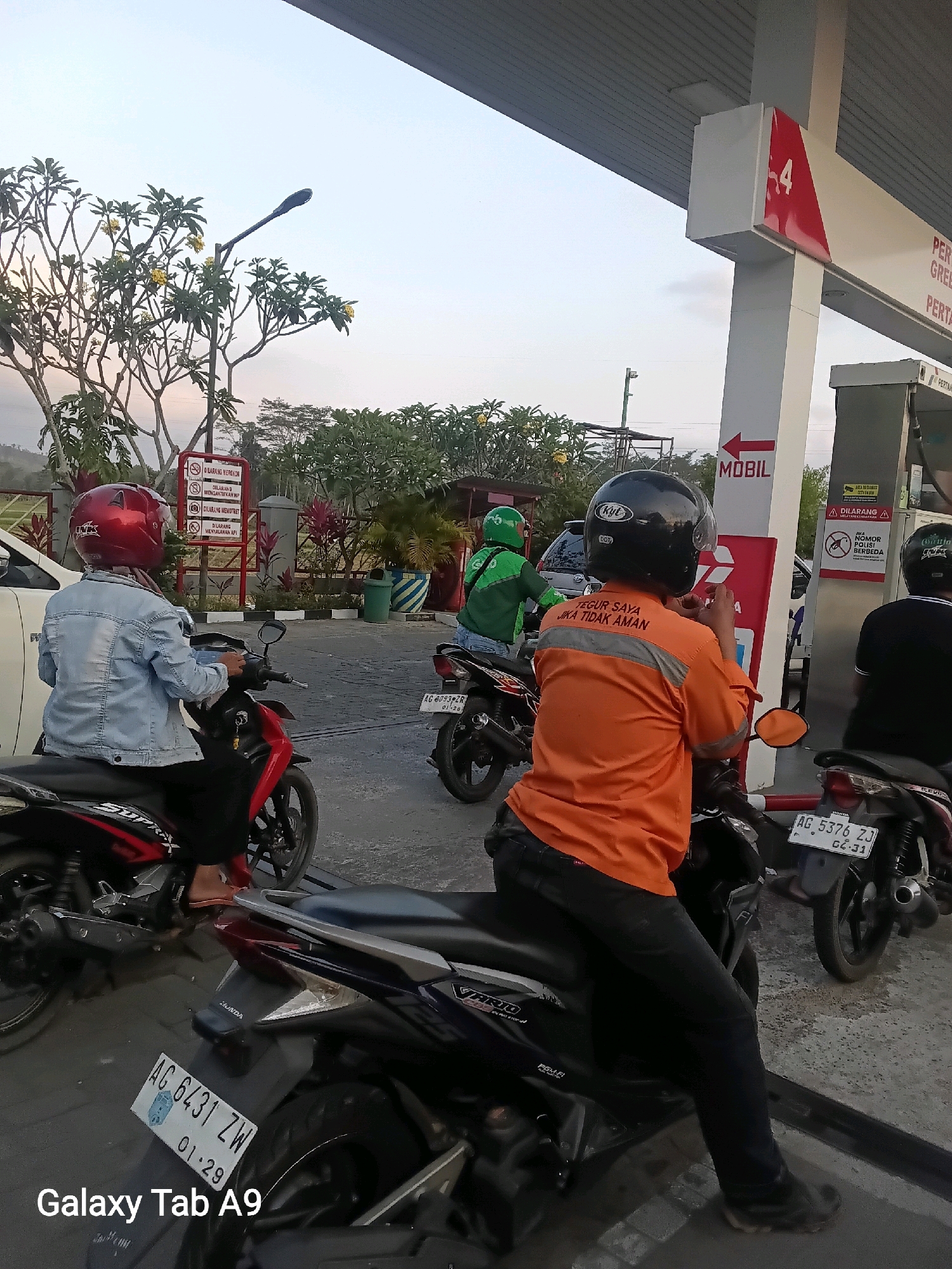The image depicts an outdoor scene at a Pertamina gas station. In the foreground, several individuals on motorcycles are visible. One person, wearing a red helmet and a denim jacket, is seated on a red and black motorcycle. Another person, dressed in an orange shirt with reflective stripes and a black helmet, is on a dark-colored motorcycle. The orange shirt has text that reads "TEGUR SAYA JIKA TIDAK AMAN," which translates to "Correct me if I'm unsafe." Further back, a person in a green jacket, likely a delivery driver for Gojek, is on a motorcycle.
The gas station features prominent red and white branding typical of Pertamina. Signs indicating "MOBIL" (car) with an arrow pointing left are visible. There are also red and white striped poles and gas pumps. In the background, lush green trees and vegetation suggest a rural or semi-rural setting, with a glimpse of hills or mountains in the distance. The sky is clear and appears to be late afternoon or early evening. The presence of the Pertamina branding, delivery driver uniforms, and typical Indonesian license plates (e.g., AG xxxxxx ZW) strongly indicate that this location is in Indonesia. Given the license plate prefix "AG," this suggests the vehicle is registered in the province of East Java, and therefore, the gas station is likely located within or near the city of Trenggalek, East Java, Indonesia.