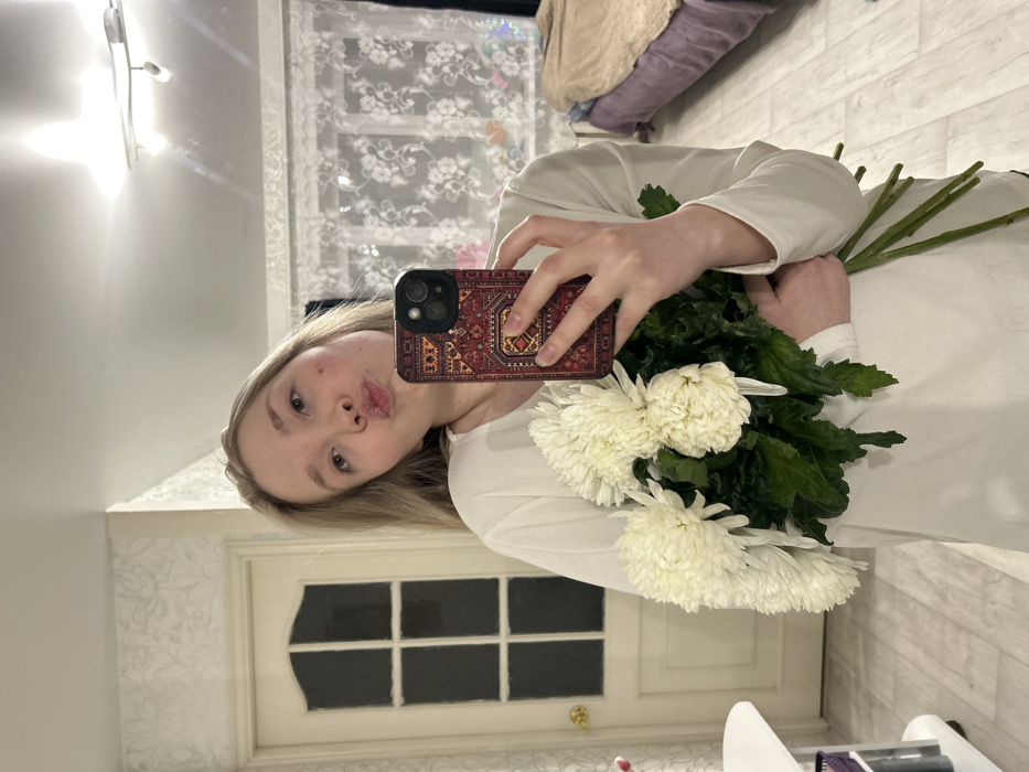 In an interior residential space in Perm, Russia, a young female with light-colored hair is capturing a mirror selfie. She wears a white, long-sleeved top and holds a dark-colored smartphone with a red, intricately patterned case in her right hand, positioned towards the mirror. Her lips are pursed, forming a "kiss" gesture, and she gazes directly at the phone's camera. In her left arm, she cradles a bouquet of approximately six large, white chrysanthemums with prominent green foliage.
The background on the left features a light-colored, paneled door with multiple glass inserts and a gold-colored doorknob. The adjacent wall is adorned with light-toned wallpaper featuring a subtle floral or vine pattern. A portion of a white surface, possibly a shelf or table, is visible in the lower-left corner.
To the right, a window is covered by white lace curtains with a distinct floral pattern. Beyond the curtains, a faint blue light source or object is discernible. Below the window, a bed is present, covered by a light brown blanket over a purple or lavender sheet. The ceiling above features a white track lighting fixture with two directional spotlights. The flooring throughout the visible area is light-toned, appearing to be laminate or tile with a wood-grain texture.