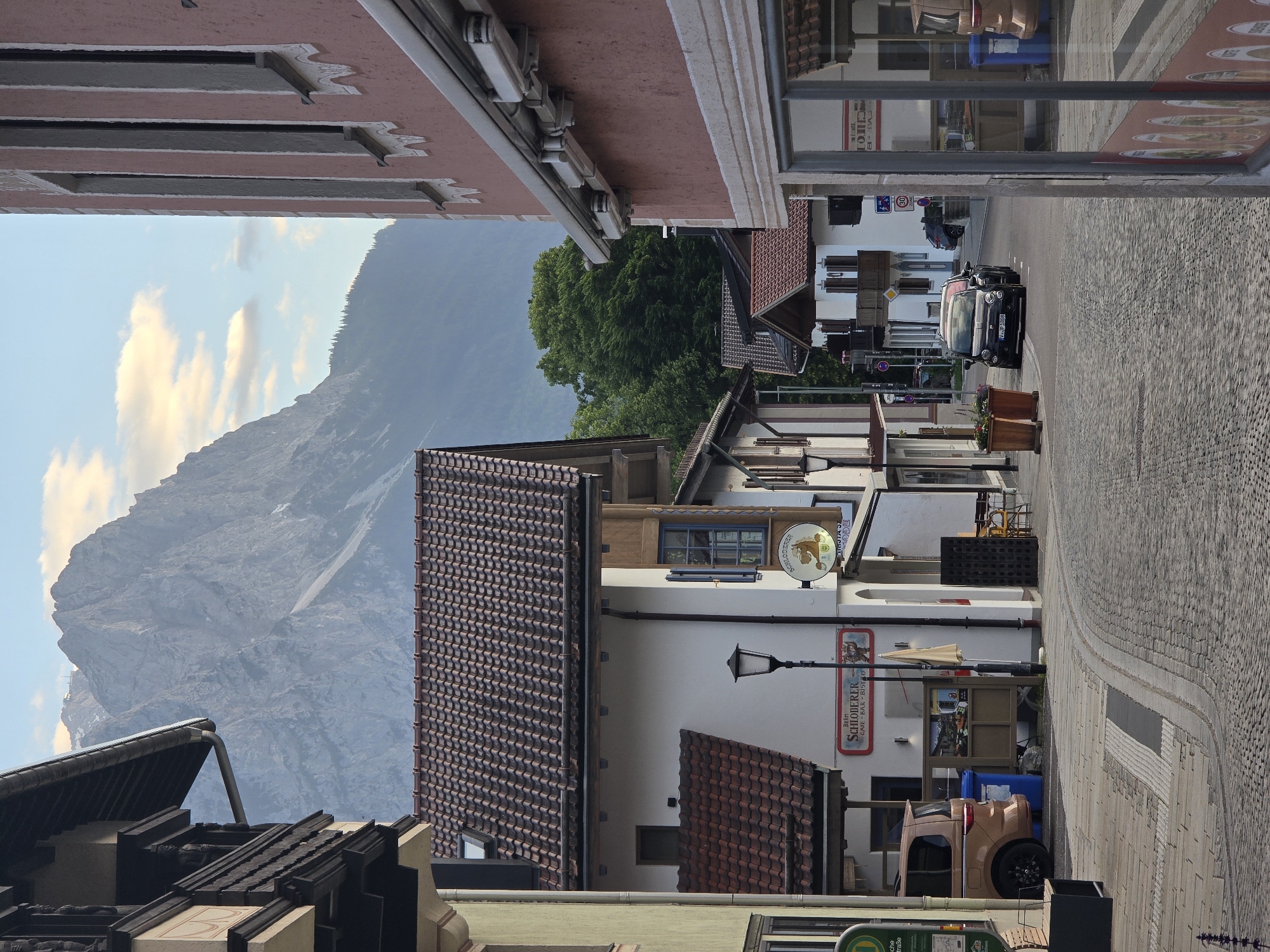 The image captures a street scene in Garmisch-Partenkirchen, Germany, with a prominent mountain range in the background. The foreground features a cobblestone street lined with buildings in a traditional Bavarian architectural style. On the left, a building with a dark wooden balcony and a tiled roof is visible. Adjacent to it is a white building with red-tiled roofs, displaying signs for "Beim Schloderer Cafe Bar Bistro." A brown minivan is parked on the left side of the street.
To the right, a building with a pink facade and white decorative trim occupies the immediate foreground, reflecting the street scene in its windows. Further down the street, a small black car is driving away from the viewer. The background is dominated by a large, imposing mountain, possibly the Zugspitze, with some clouds around its peak. Lush green trees flank the street and the base of the mountain. The sky is partly cloudy with patches of blue. The overall impression is of a charming, picturesque alpine town.