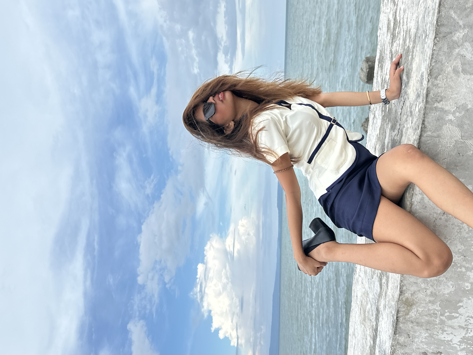 The image depicts a young woman with long, brown hair and sunglasses in a dynamic pose, stretching her leg while sitting on a concrete ledge by the sea. She is wearing a white top with navy accents and a navy skirt. The background features the ocean and a cloudy sky, suggesting an outdoor setting in a coastal area. The specific location is identified as Roxas Avenue in Davao City, Philippines, a prominent street known for its seaside promenade. The overall scene captures a moment of leisure and physical activity against a scenic backdrop.