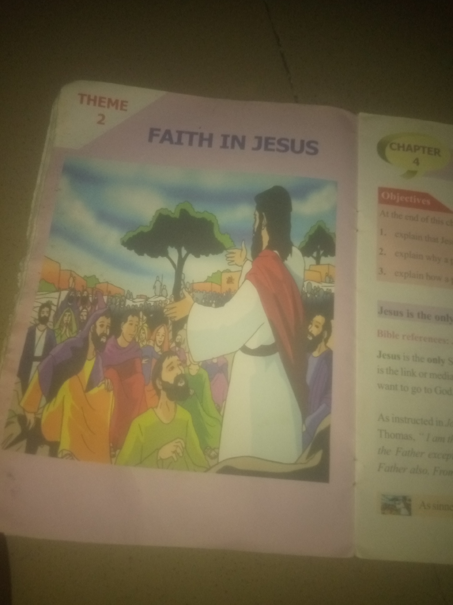 The image shows an open book with illustrations and text. The left page features a colorful illustration of Jesus speaking to a crowd of people in a stylized, cartoonish manner. The people are depicted in various clothing and expressions, with some appearing to be listening intently. In the background, there are buildings and trees. The text on the left page includes "THEME 2" and "FAITH IN JESUS".
The right page has a section titled "CHAPTER 4" and "Objectives". Below the objectives, there is text discussing "Jesus is the only..." and "Bible references". Further down, there is text that appears to be a quote or scripture, mentioning "Thomas", "the Father", and "Father also". There is also a small image of a Nativity scene.
The overall scene depicts religious educational material. The image does not contain any recognizable elements that would indicate a specific location within the city of Jauro Yinu, Nigeria. The environment depicted is an illustration and not a photograph of a real place.