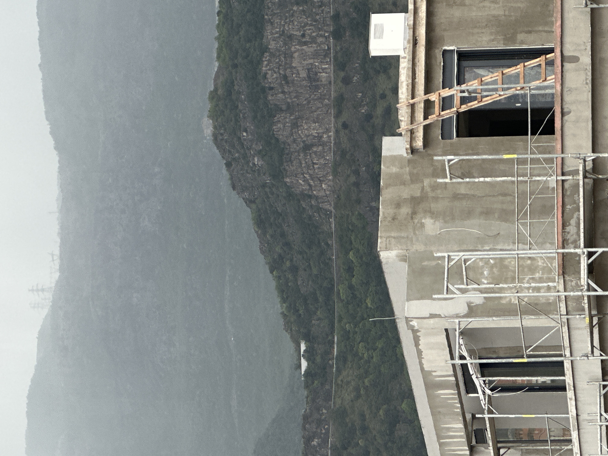 The image displays a multi-story concrete building under construction, viewed from a slightly elevated position, with a vast mountainous landscape in the background. The building, occupying the right side of the frame, is unfinished, showing exposed concrete walls and several window openings without glass. Scaffolding made of metal poles and platforms is erected along its facade, suggesting ongoing work. A wooden, ladder-like structure is precariously propped against one of the upper window openings. A small, white rectangular outdoor unit, possibly an air conditioning unit, is mounted high on the building's exterior. Electrical conduits and wires are visible in various places on the unfinished walls.
In the mid-ground and background, densely forested mountains rise, characterized by rugged, rocky sections and lush green vegetation. A narrow road or path can be discerned snaking along the lower slopes of the mountains. The furthest mountains are shrouded in atmospheric haze, indicative of an overcast sky or misty conditions. The overall lighting is soft and diffuse, suggesting a cloudy day. No people are visible, but the scaffolding and unfinished state imply active construction. There are faint yellow strips on the scaffolding; one close-up shows what appears to be unreadable text or a logo.
