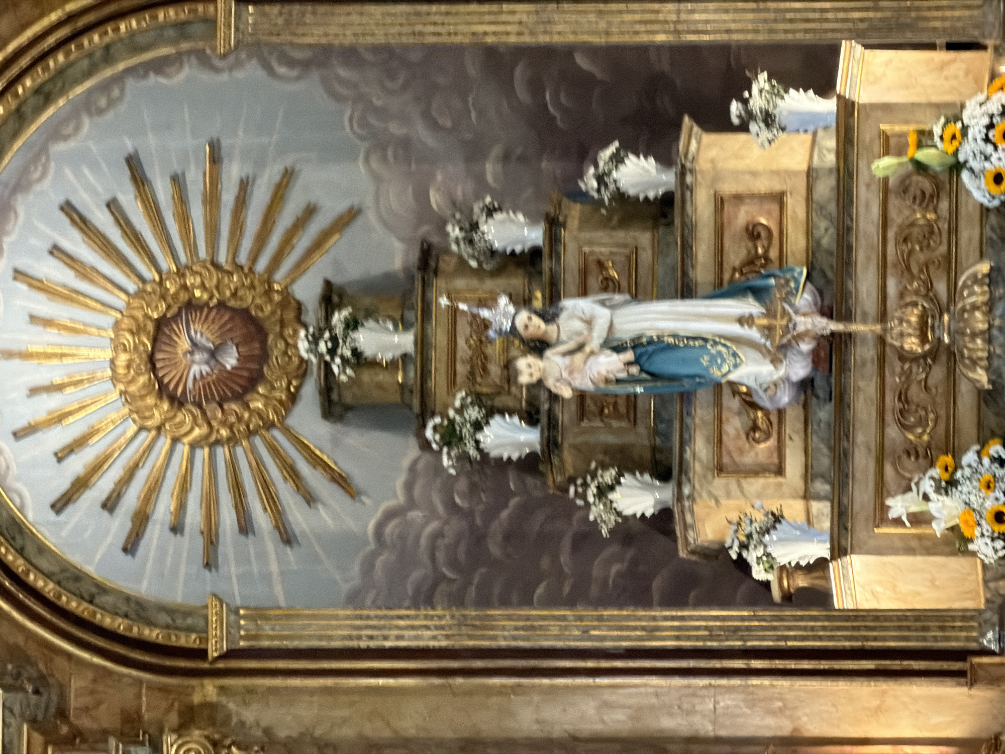 A close-up, slightly rotated, full-shot showcases the altar of the Capela de São João in Vila Real, Portugal. At the center of the shot is an arched painting that contains a gold-rimmed sun with golden sunbeams. The sunbeams emanate from a circular golden fixture with an image of a dove. Below the painting, in the middle right of the shot, are various items, including a statue of a woman in blue and white robes. She is holding a baby and standing on a gold pillar. The statue is surrounded by flower arrangements. The backdrop of the altar is a painted sky with clouds.