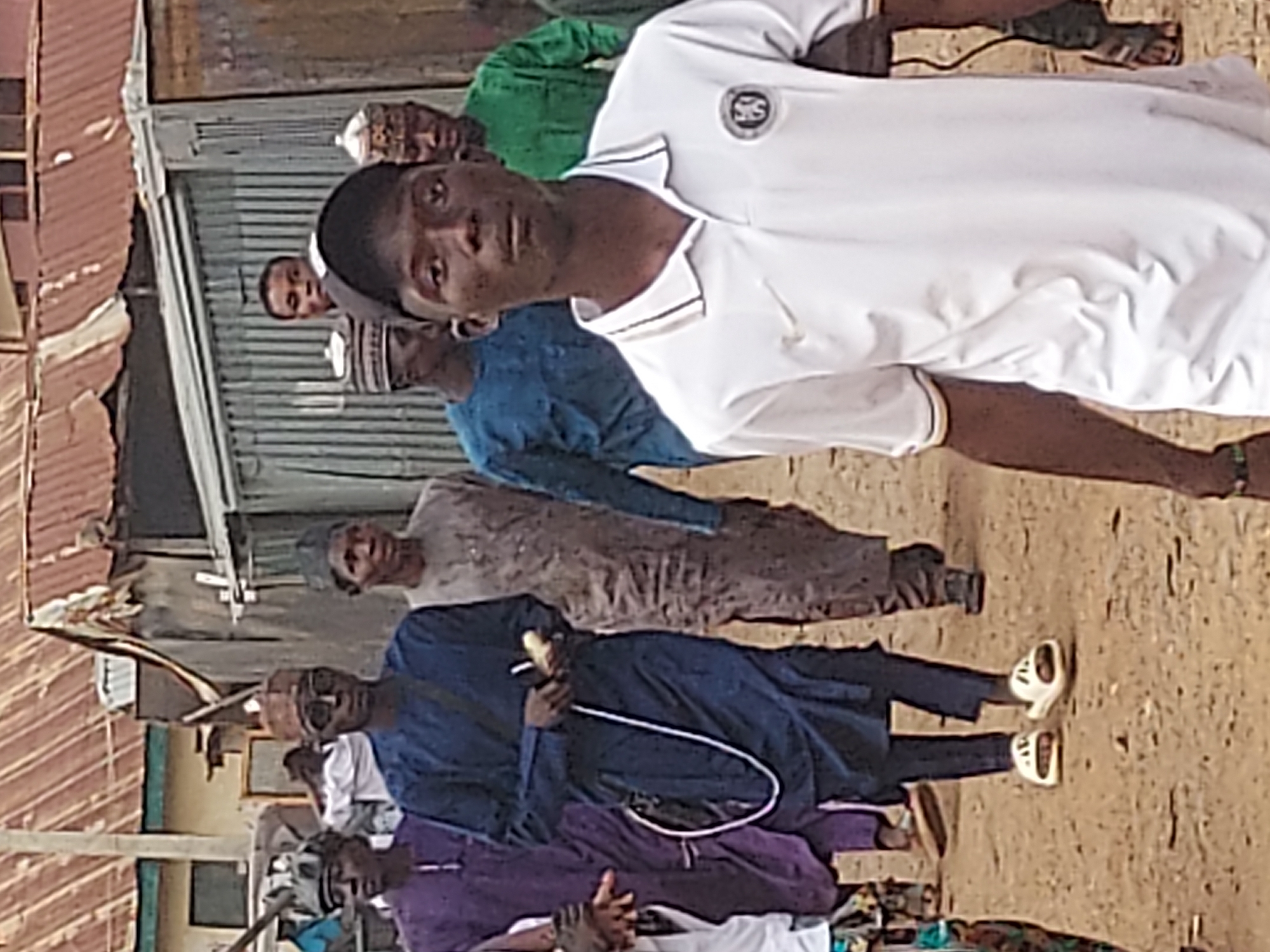 In an outdoor setting, likely a street scene in Jos, Nigeria, a group of people is captured. The focal point is a young man in the foreground, wearing a white shirt with a small logo on the upper left chest, and dark shorts, looking directly at the camera. Behind him are several other individuals of varying ages and attire, some wearing traditional Muslim caps and robes in colors such as blue, green, and brown. The environment includes buildings with corrugated metal roofs in the background. The ground appears to be a sandy or dirt surface. One man is holding a long pipe. Several are wearing shoes or sandals. The people appear to be gathered in a line or group, suggesting a possible gathering or procession.