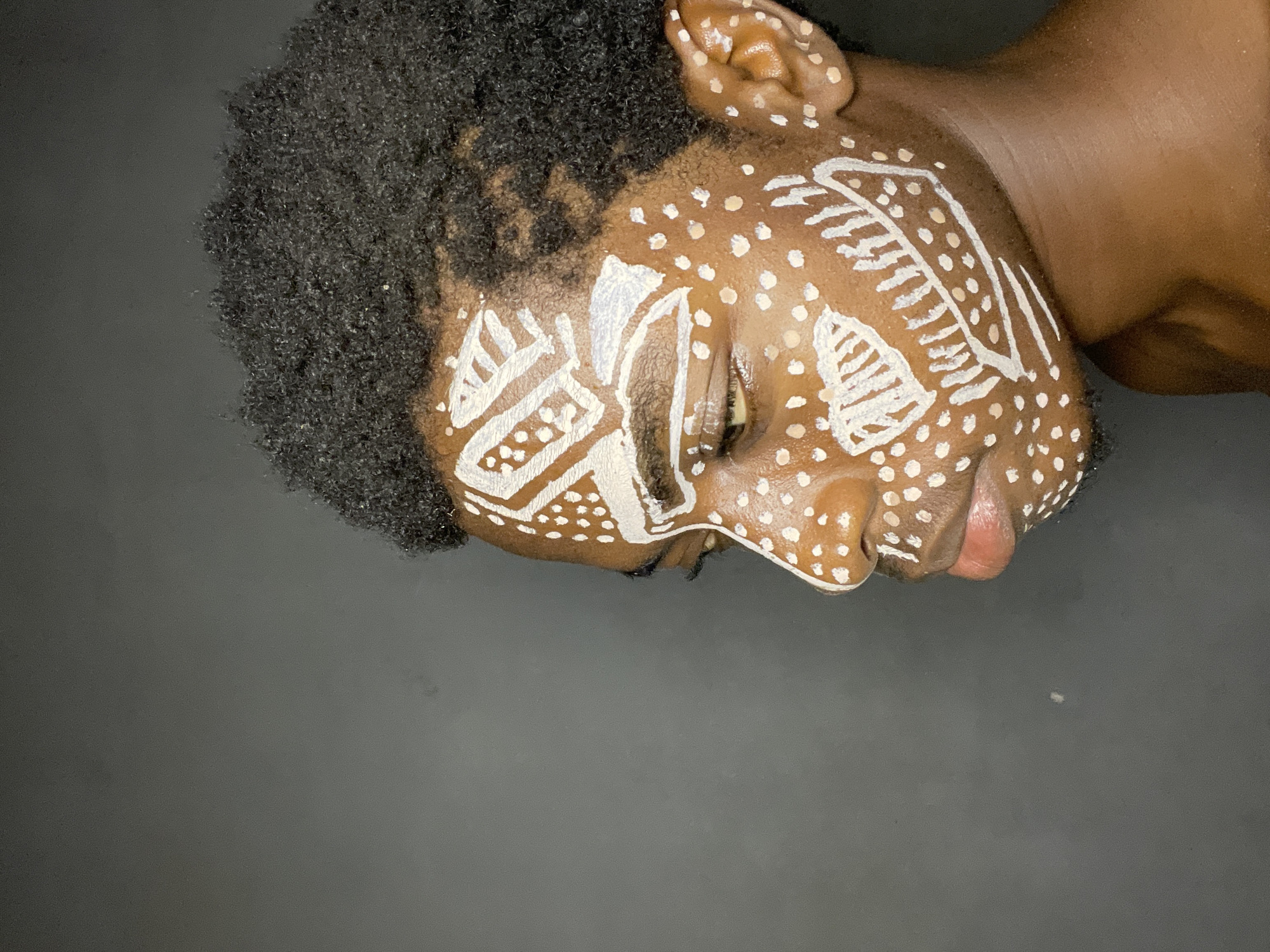 A medium shot shows a person's face, adorned with white paint tribal markings, against a dark gray background. The person has dark skin and tightly curled black hair. The white paint is applied in patterns including dots, lines, and geometric shapes across the forehead, cheek, and ear. The person's eyes are closed. The setting is presumably a studio or controlled environment given the background. There is no information to place the image in Nkpor, Nigeria.