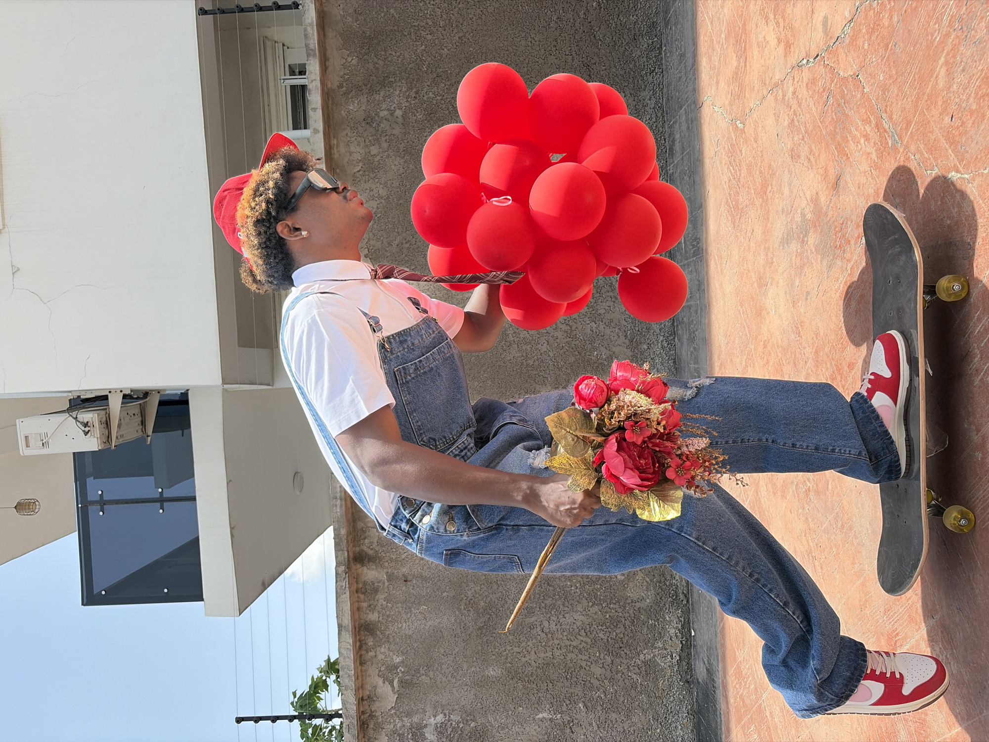 A young man stands on a skateboard in Igboefon, Nigeria, wearing red and white sneakers, denim overalls, a white shirt with a patterned tie, and sunglasses. He also wears a red hat. He is holding a large bouquet of red balloons and a bouquet of red and gold artificial flowers. The background consists of a building with a window, a concrete wall, and a portion of the blue sky. The scene is captured in daylight.