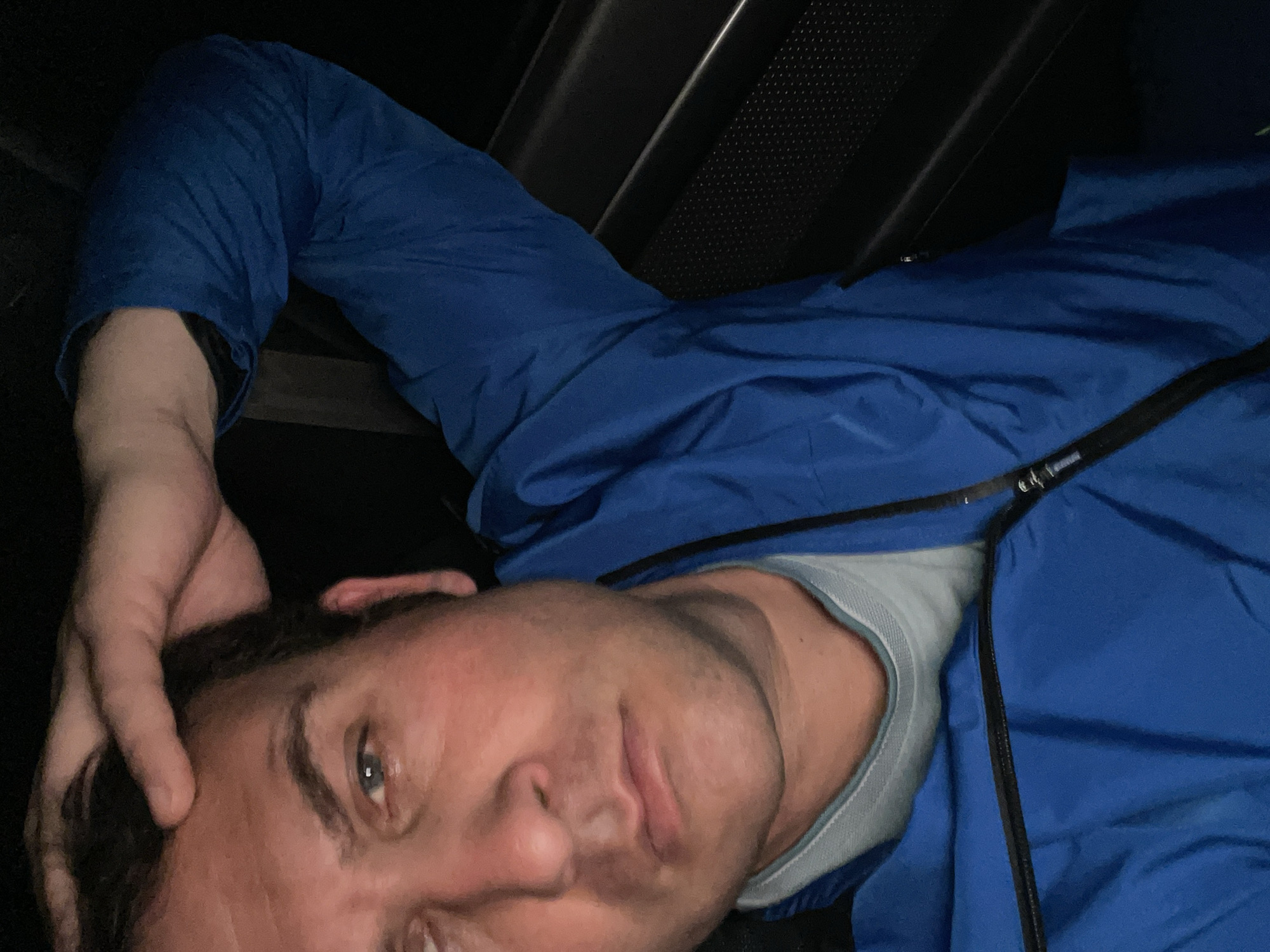 A man is depicted lying down in a dark, enclosed space, possibly within a vehicle, in Armagh, United Kingdom. He is wearing a blue zip-up jacket over a light blue or grey crew-neck shirt. His right hand is placed behind his head and neck, suggesting he is resting or relaxing. He looks directly at the camera with a neutral to slightly relaxed expression. The background is dark, featuring angular, textured surfaces that resemble interior paneling, possibly including a speaker grille. The lighting is dim, indicating an indoor setting or nighttime. There is no visible text.