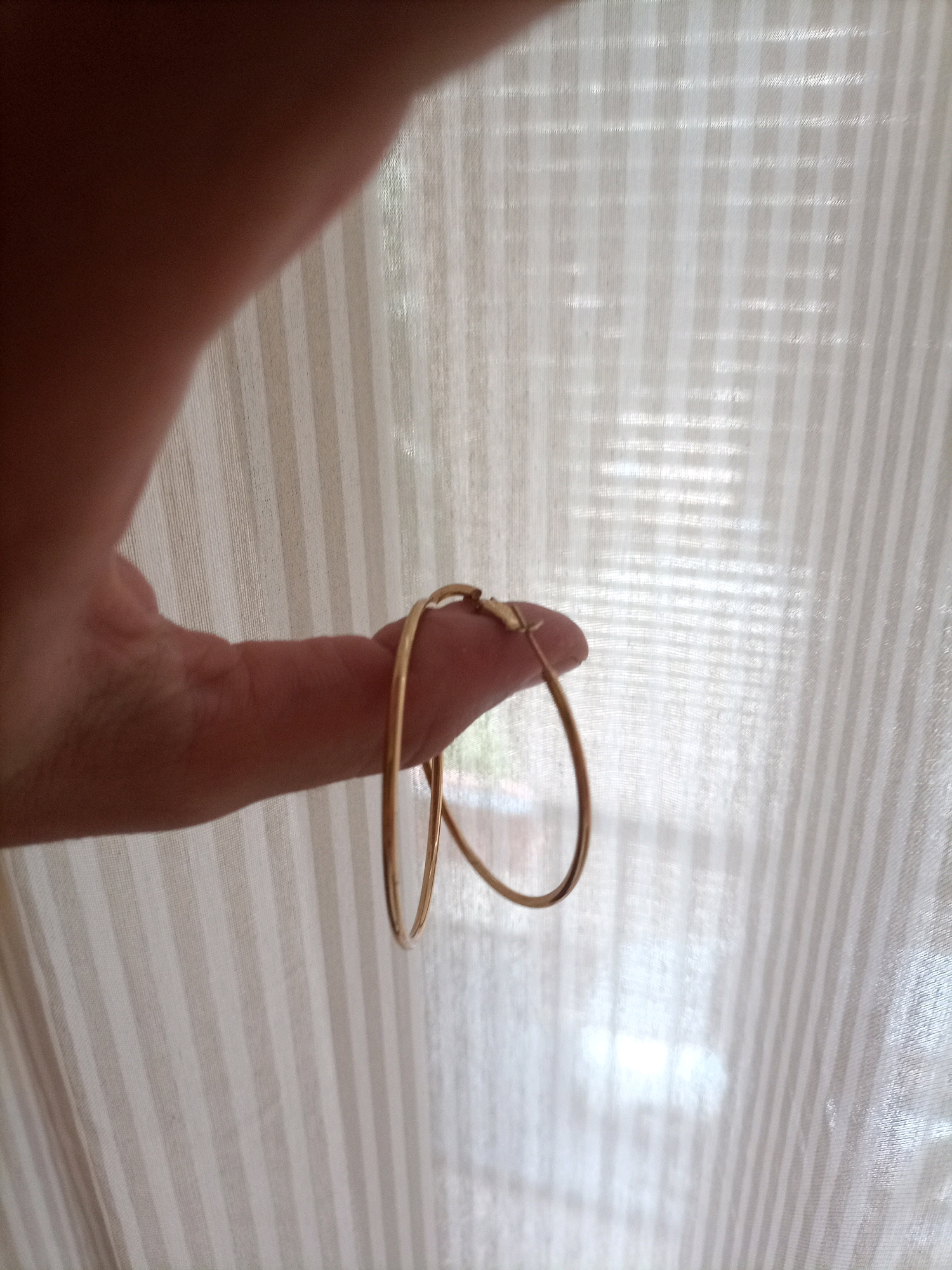 A person's thumb and index finger are holding a pair of gold hoop earrings. The background is a white, vertically striped fabric, likely a curtain or blind, which is translucent and allows light to pass through. The lighting suggests it is daytime. The image does not provide any information about the city of Tarragona, Spain.